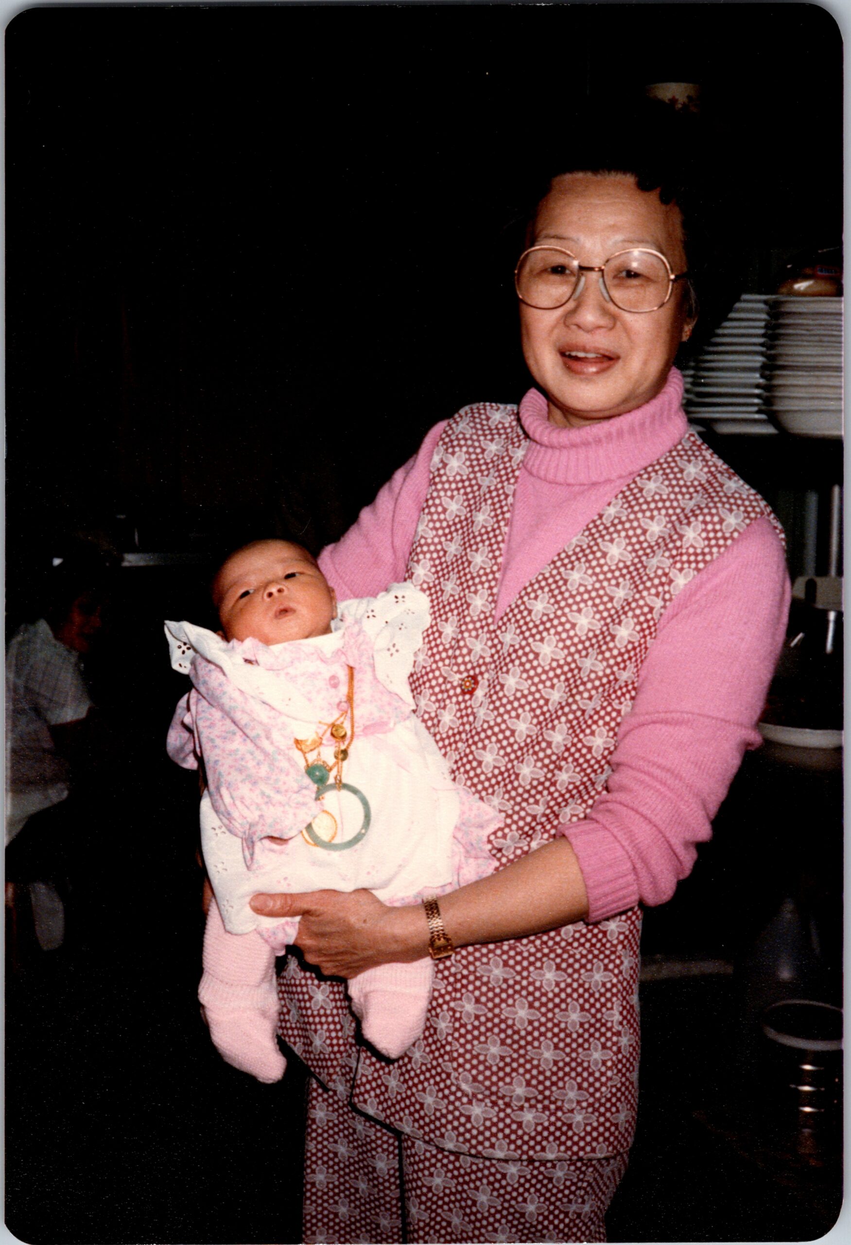 Smiling older woman with round glasses holding a newborn baby wrapped in a pink and white blanket.