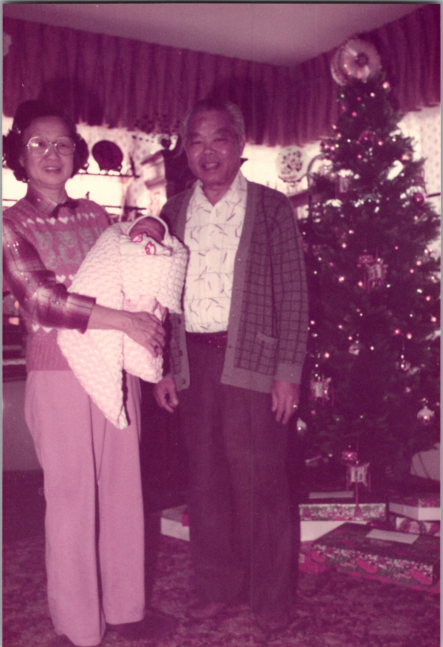 Older couple posing with their newborn baby wrapped in a blanket, beside a decorated Christmas tree and gifts in a cozy living room.