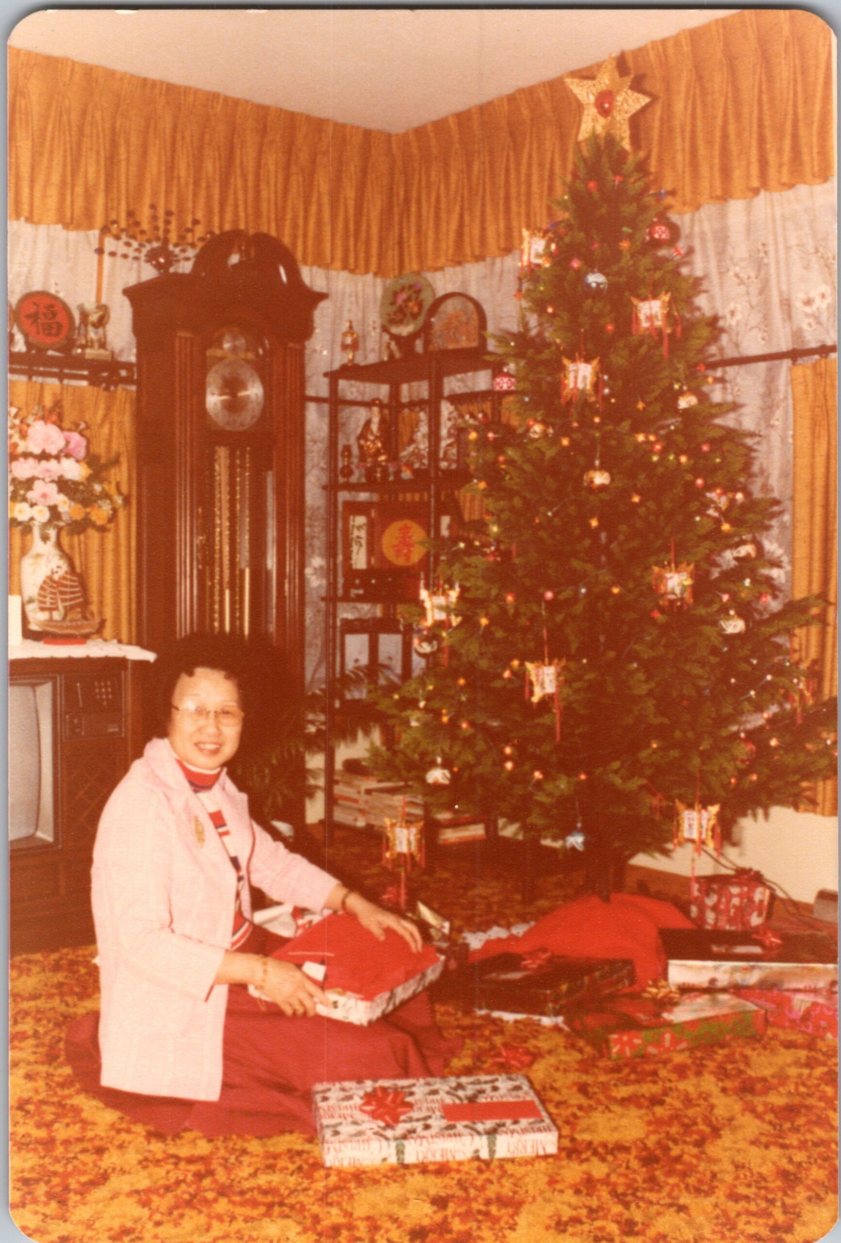 Woman in a pink jacket sits at a table wrapping a gift beside a large Christmas tree in a vintage living room.