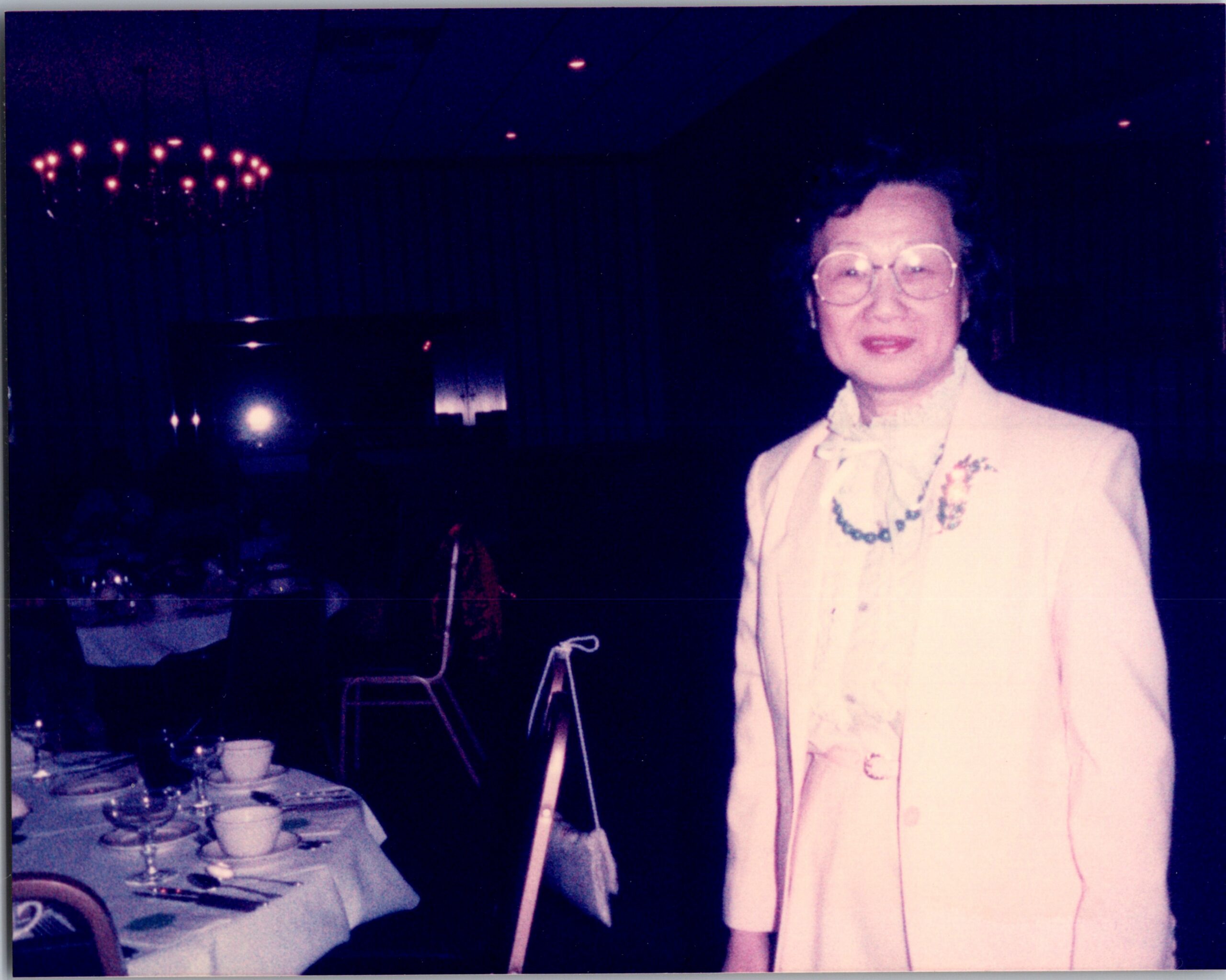 Woman in a light-colored suit and large glasses standing at a formal dinner table, smiling at the camera, chandeliers visible in the background.