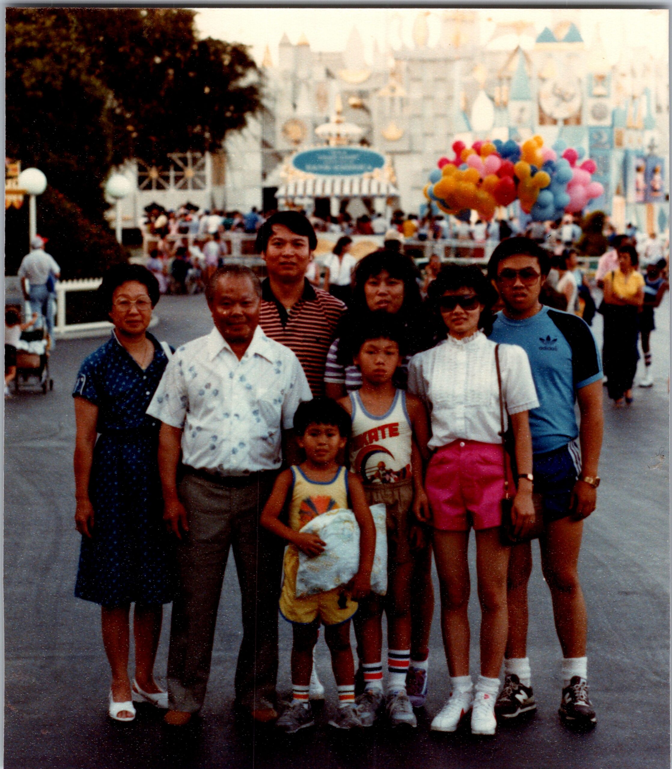 Group of adults and children posing for a photo in a busy theme park, with a pastel castle and a cluster of colorful balloons in the background.