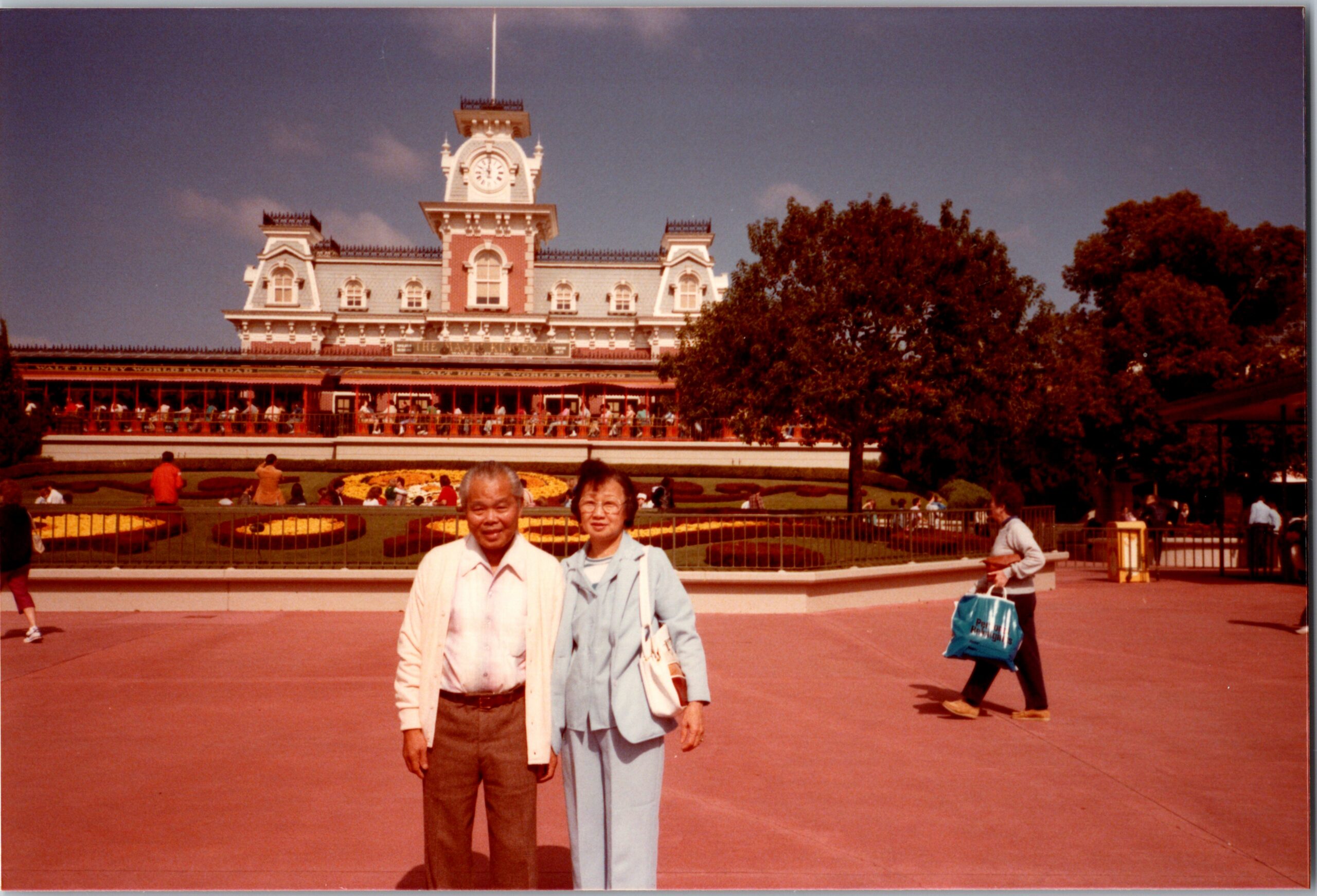 Two people pose for a photo in front of an ornate clock-tower building with a landscaped garden and trees behind them.