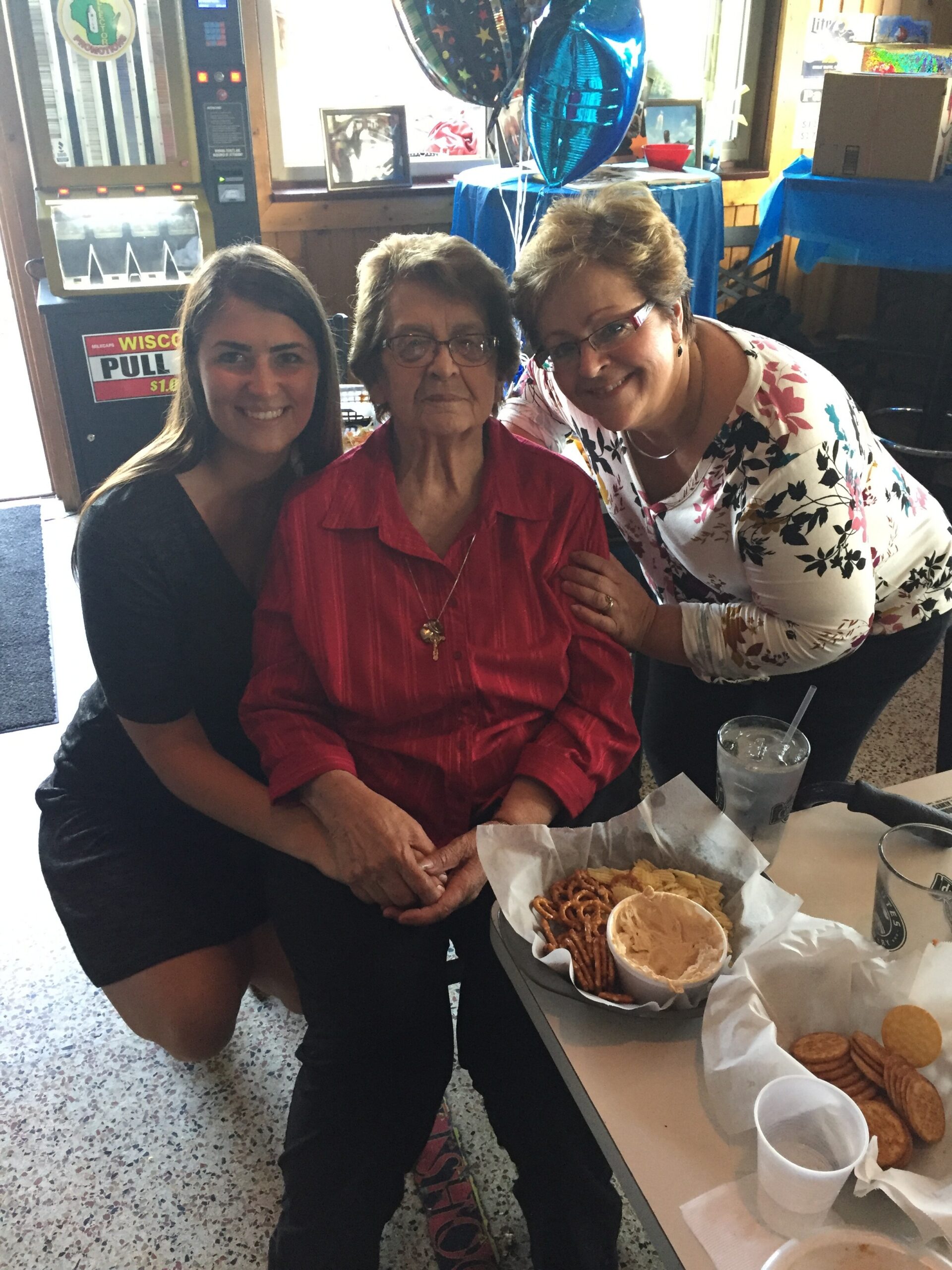 Three women pose together at a celebration table with snacks (pretzels and dip) and balloons in the background.