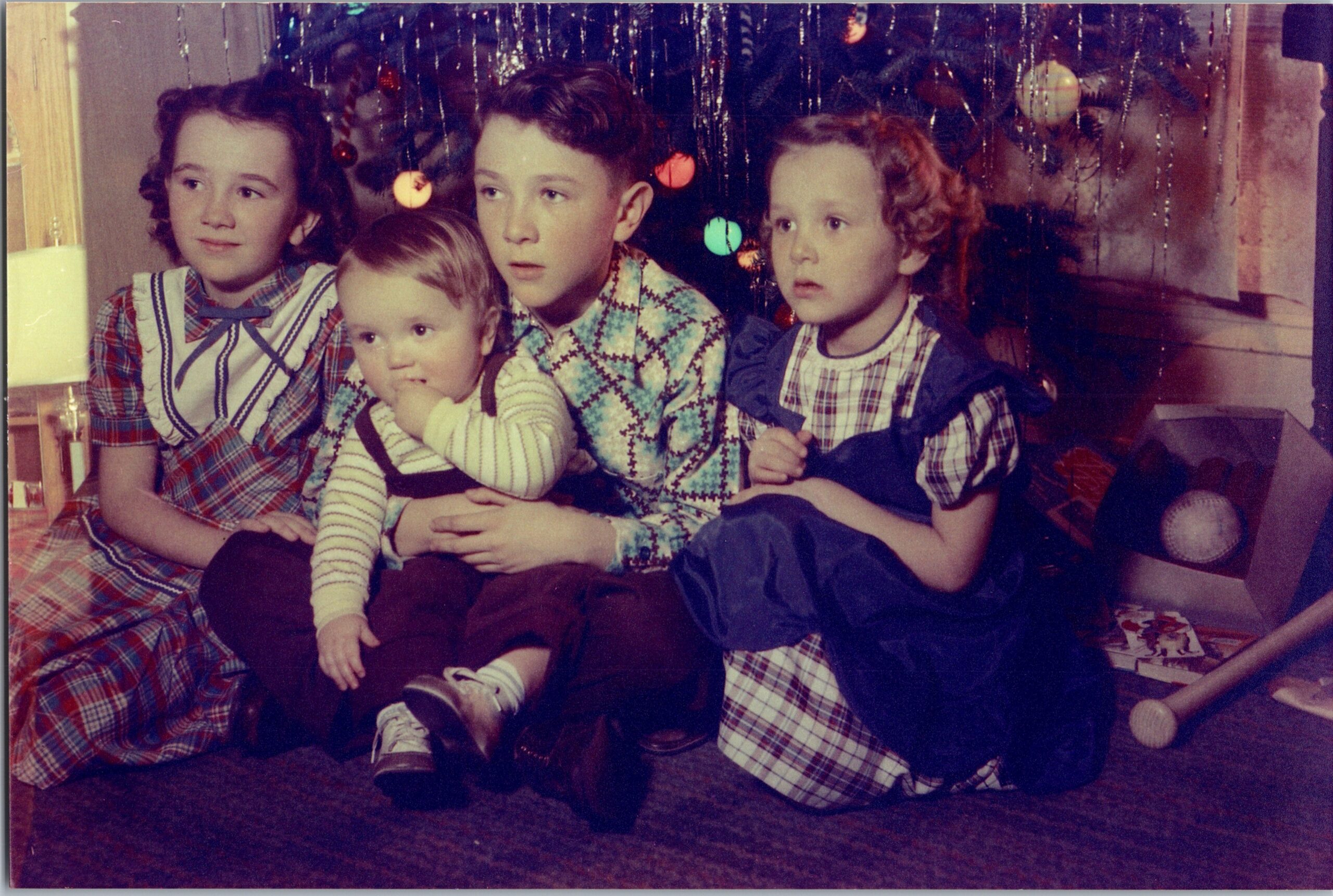 Four children sit together on the floor in front of a decorated Christmas tree indoor, dressed in plaid outfits and sweaters, looking off-camera.