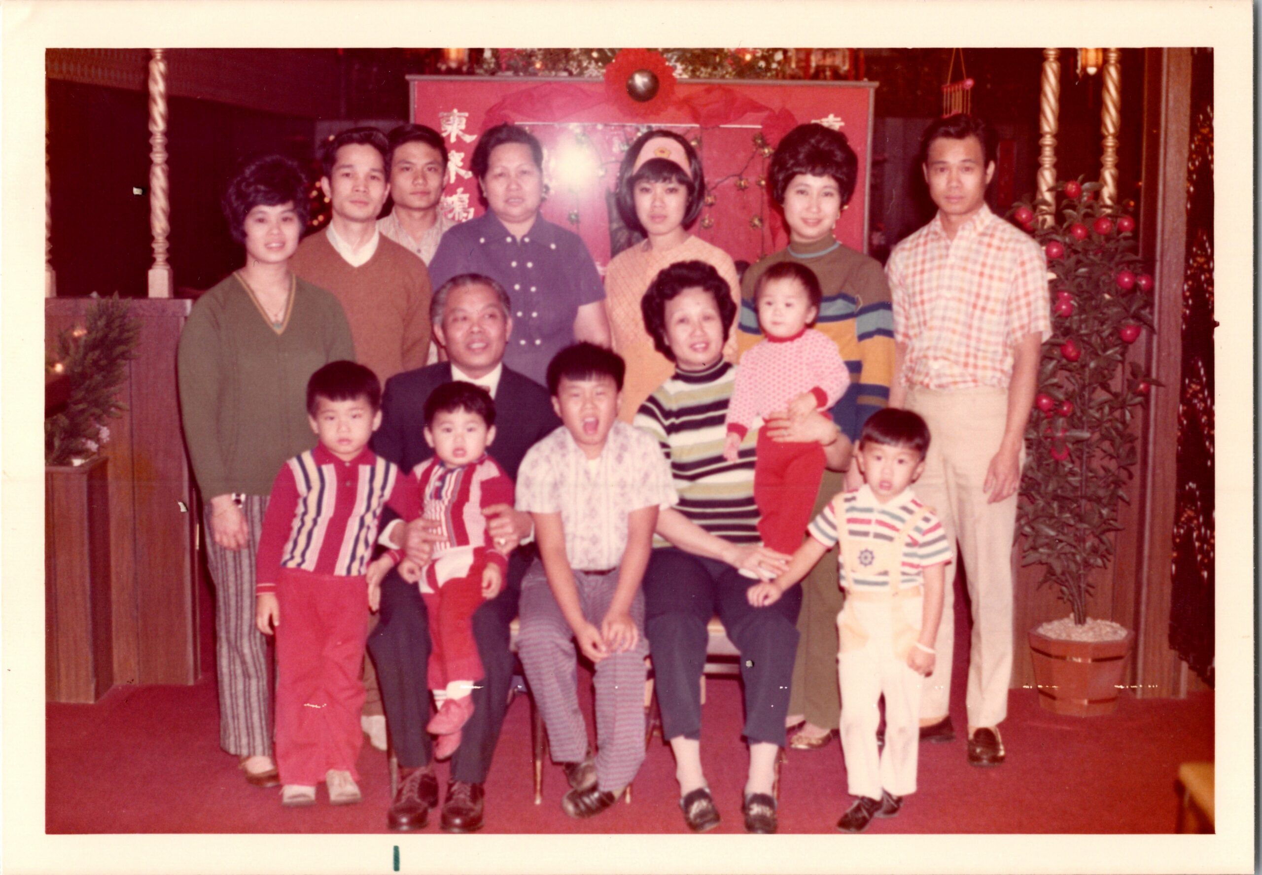 A multi-generational family posing together indoors during a festive celebration, with adults and several children in colorful outfits in front of a red decorative backdrop.