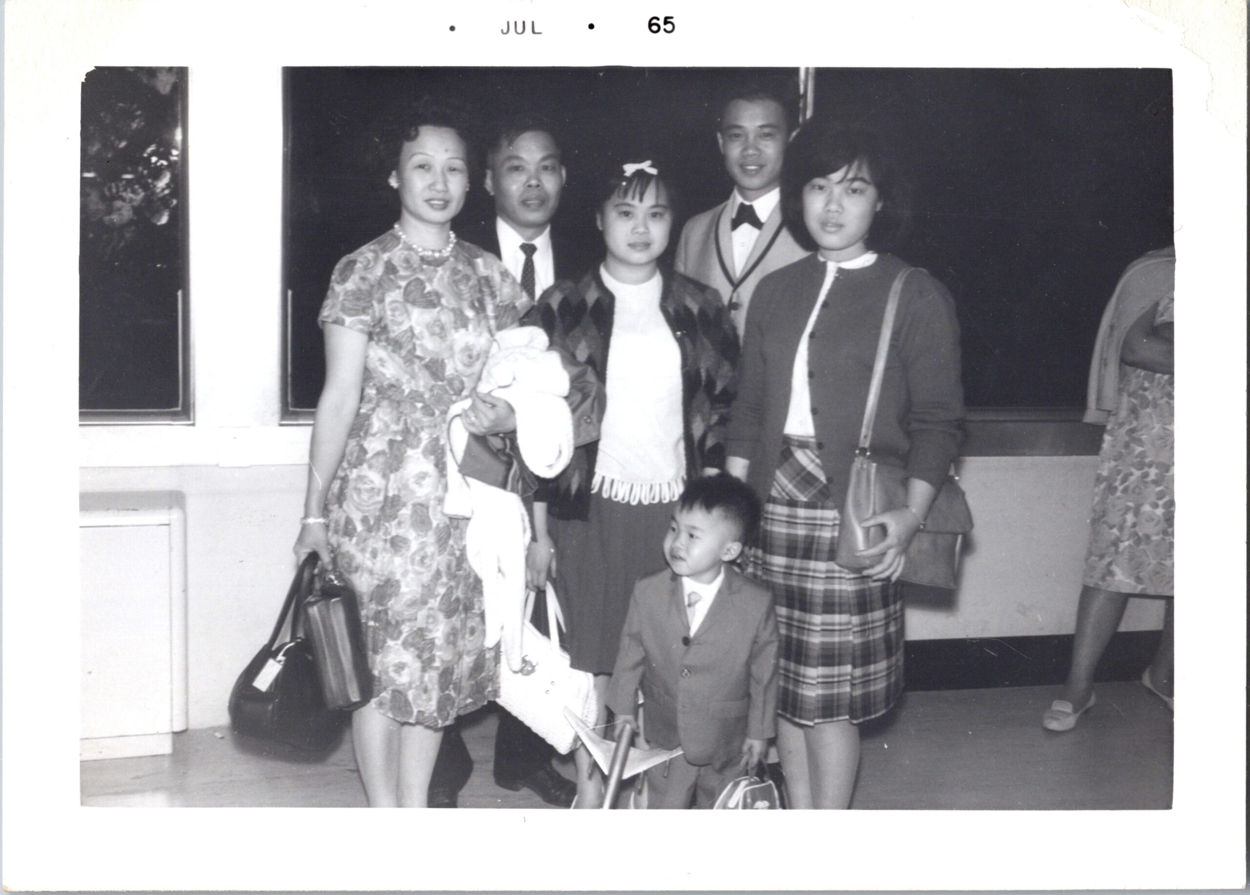 Group of five adults and two children posing together indoors in formal clothing; the woman in a floral dress stands left, others wear suits or plaid skirts, with a small boy at front.