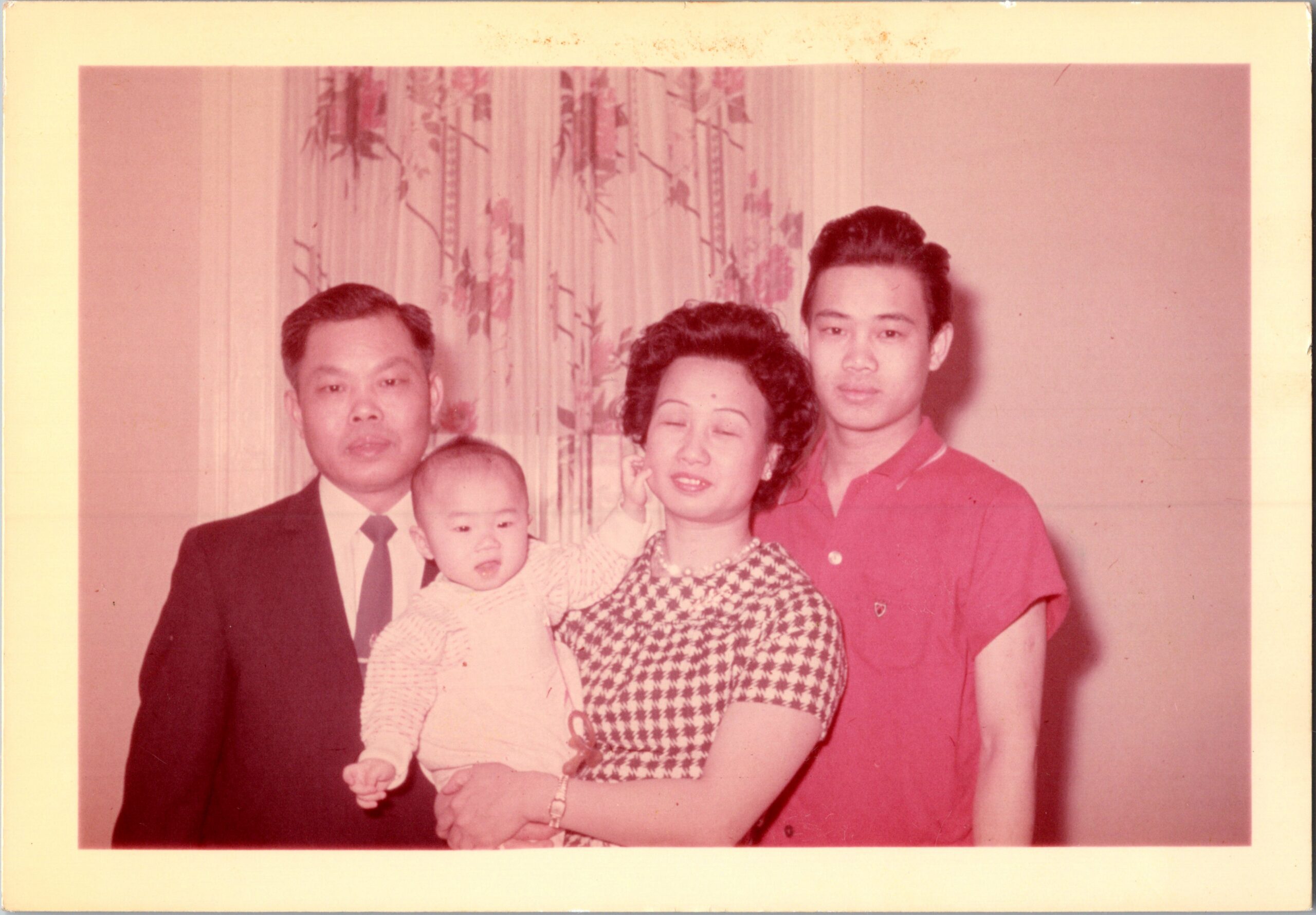 Family portrait of four adults and two babies indoors; mother in a checkered dress holds a baby, with a man in a suit on the left and a young man in red on the right.