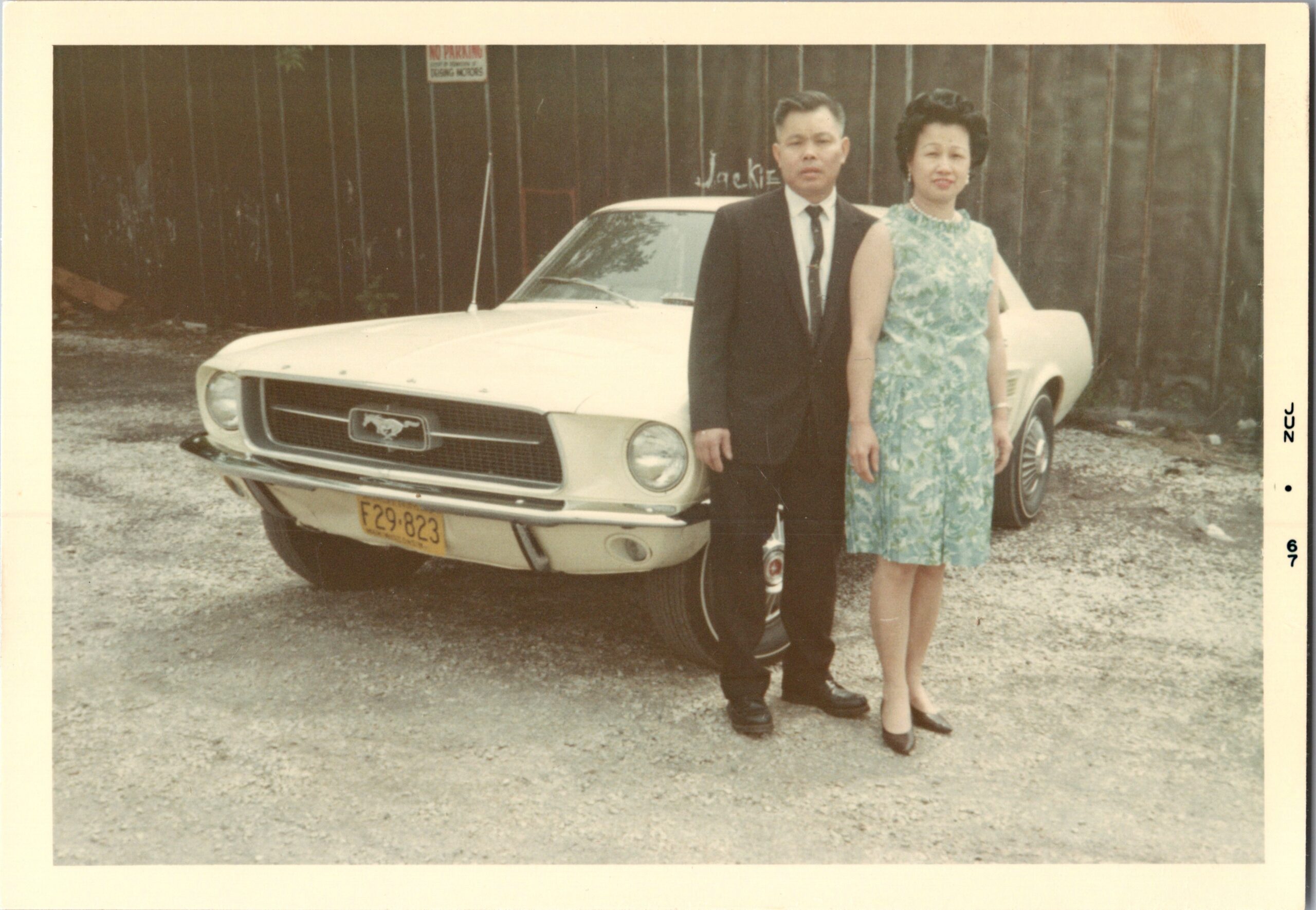 A man in a dark suit and a woman in a teal dress stand beside a cream vintage Ford, posing in a gravel lot with a wooden fence in the background.