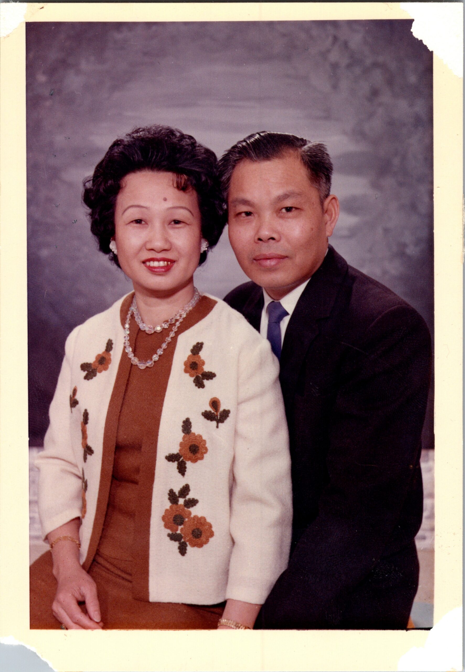 Smiling couple posing in a studio portrait: woman in a white embroidered cardigan with a brown dress and pearl jewelry, beside a man in a dark suit and blue tie.