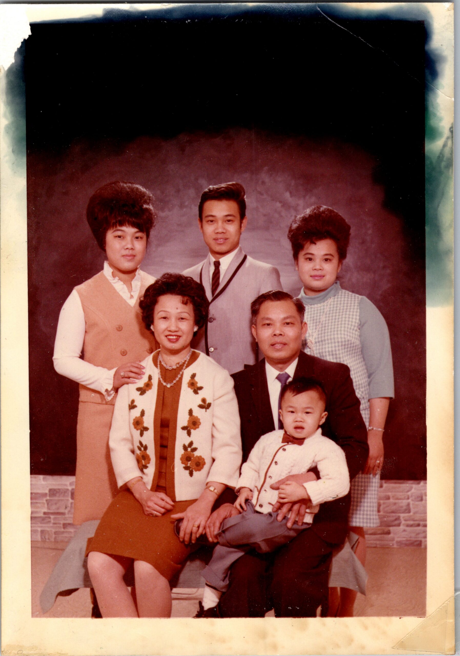 A multi-generation family posed for a studio portrait: adults standing and seated, with a baby in a cardigan front right.