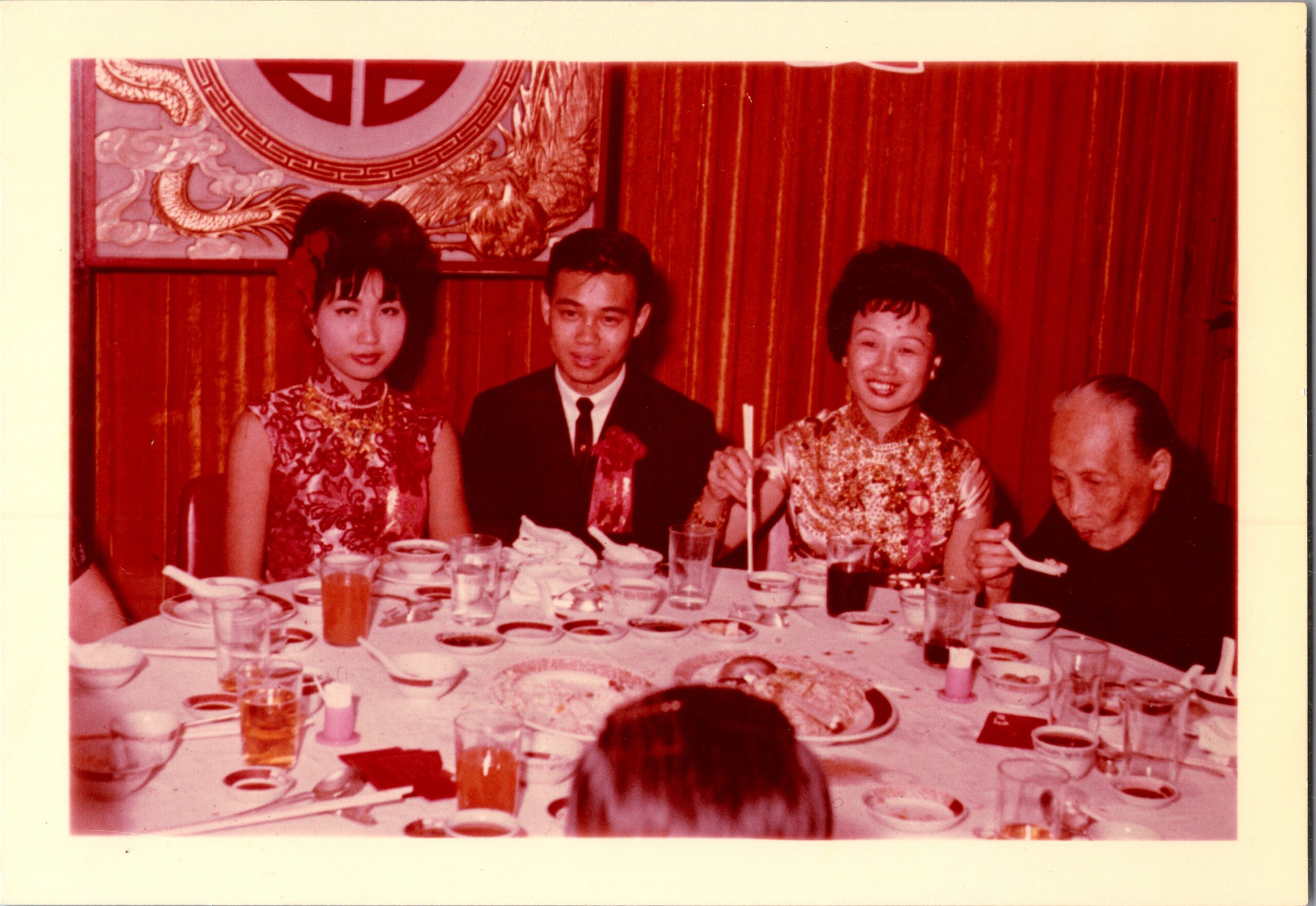 Four guests in traditional Chinese attire seated at a banquet table during a festive celebration, with red wall decor behind.
