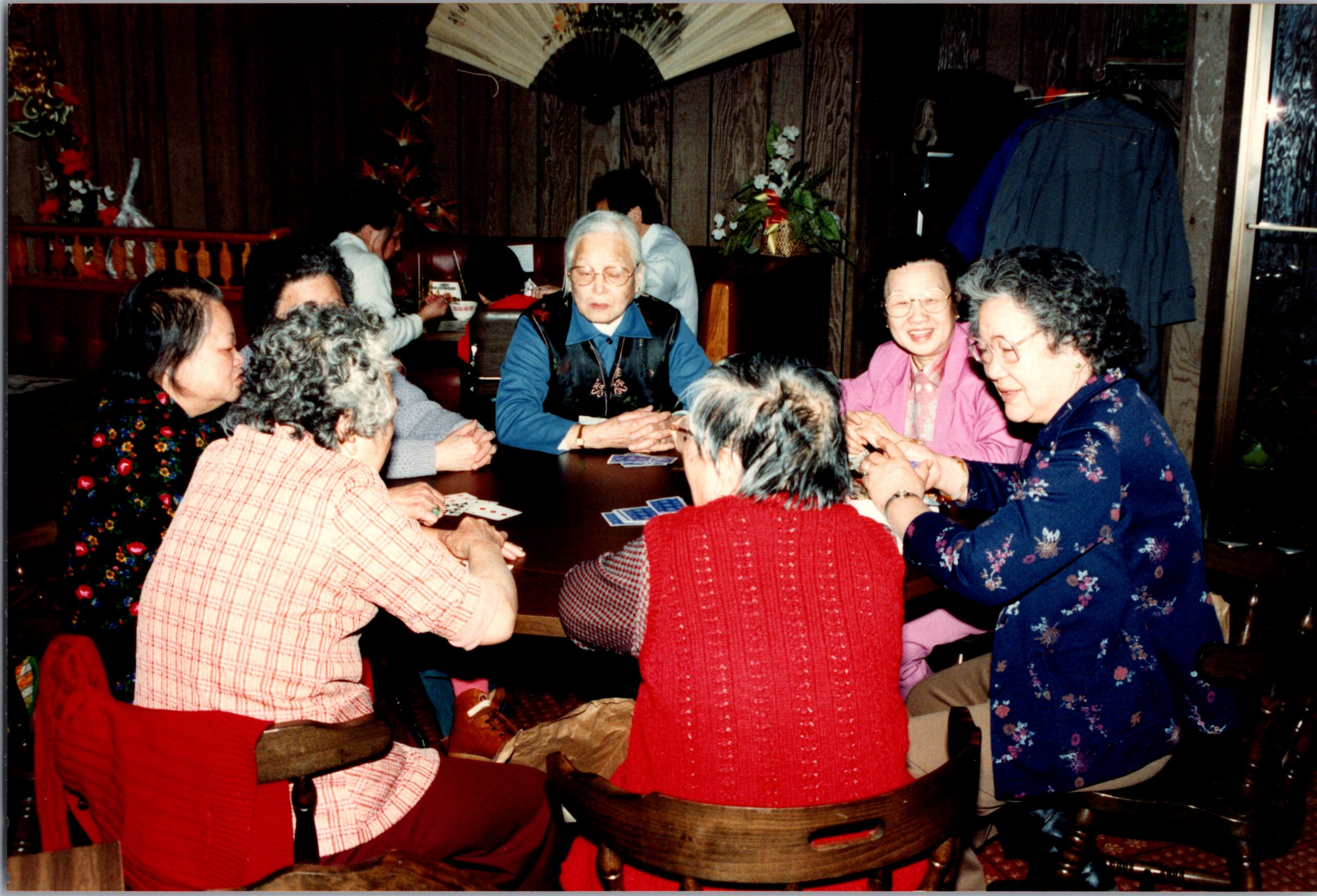 Group of elderly women gathered around a wooden table, playing cards in a wood-paneled restaurant.