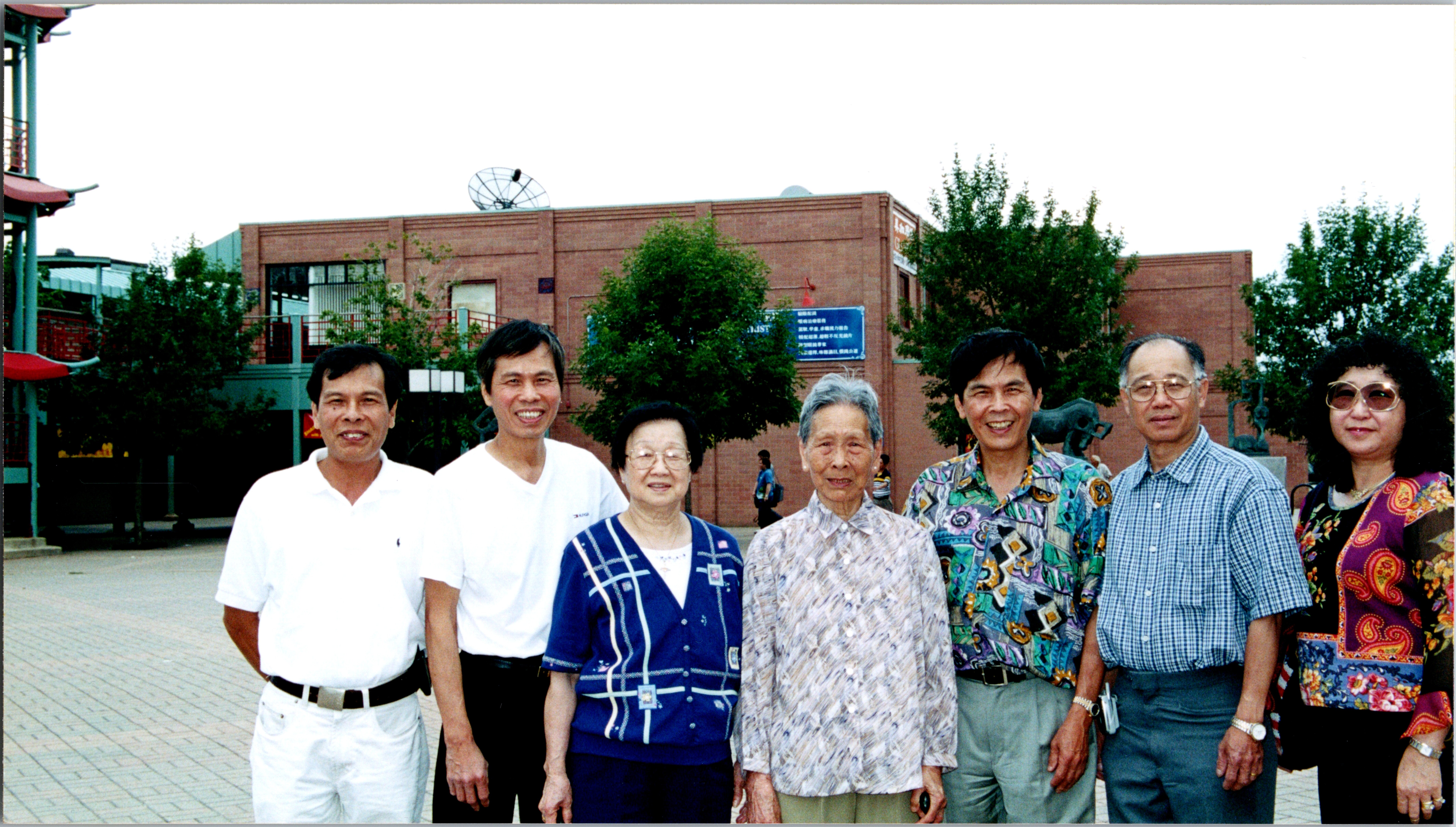 Seven adults stand together outside a brick building with trees in the background, smiling at the camera.