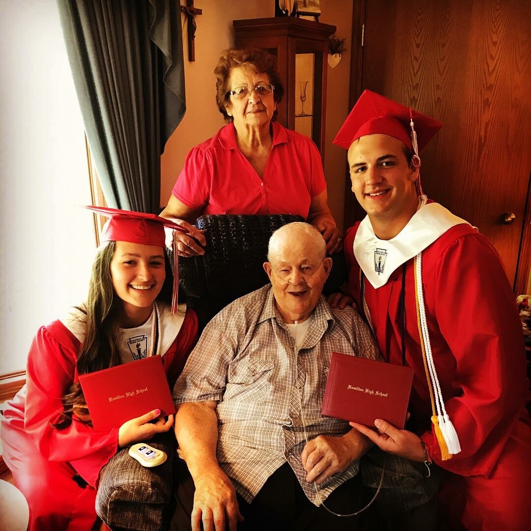 Two graduates in red cap and gown celebrate with an elderly man and a woman in a cozy home setting, each holding red diploma folders beside him.