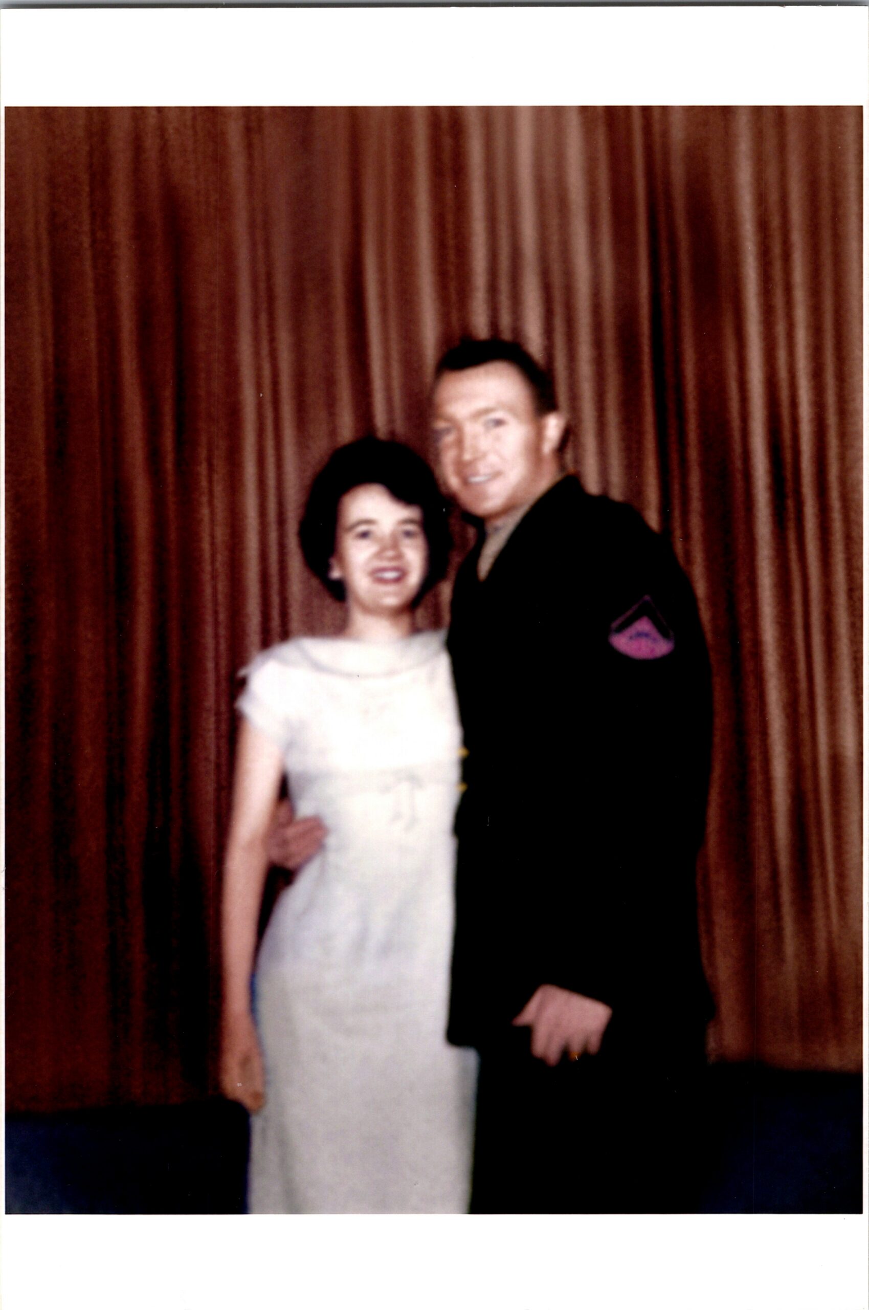 A couple posing together in front of brown curtains; woman in a light-colored dress and man in a dark uniform.