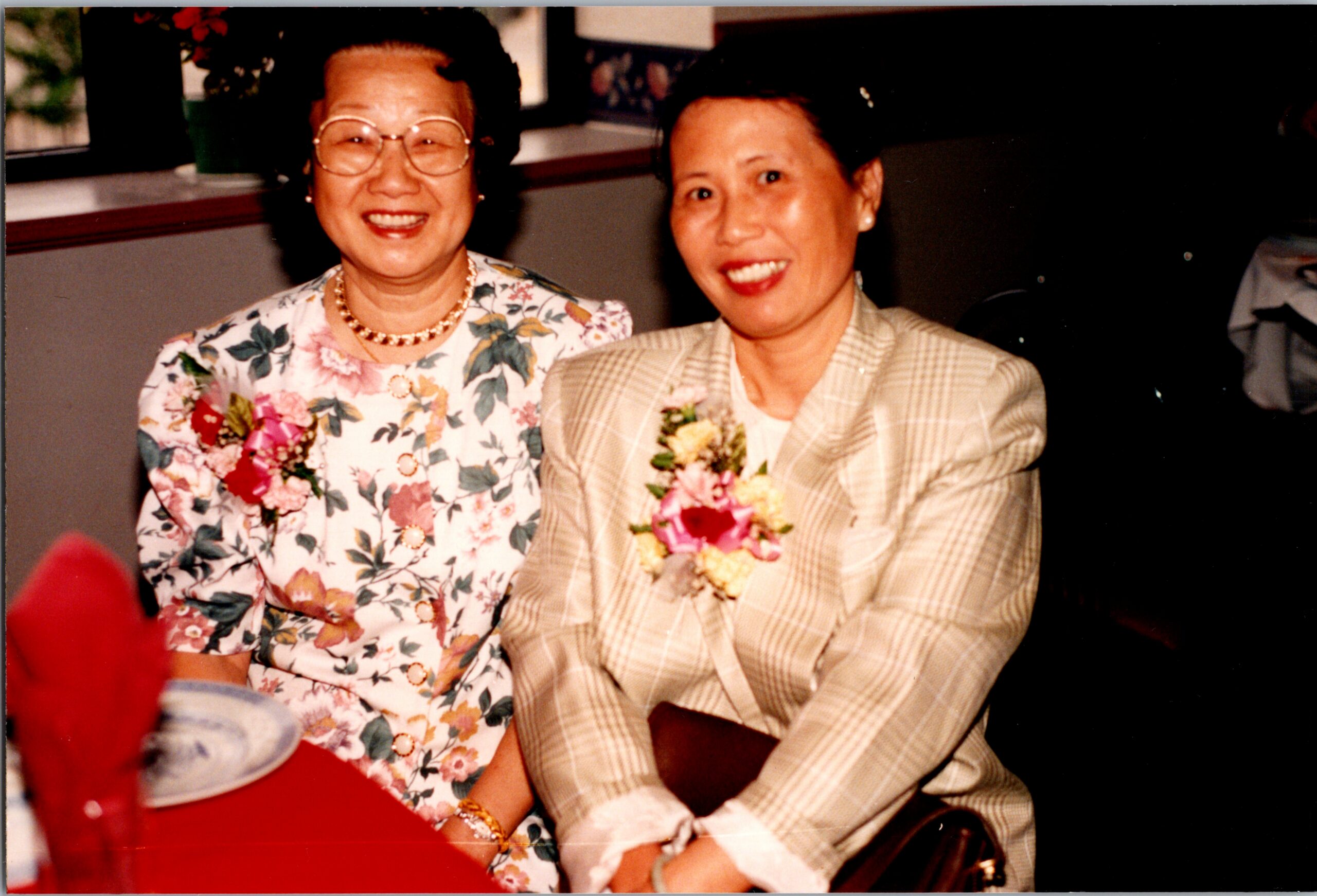 Two smiling women seated at a table during a celebration; one in a floral dress with glasses and pearl necklace, the other in a beige plaid blazer with a corsage.