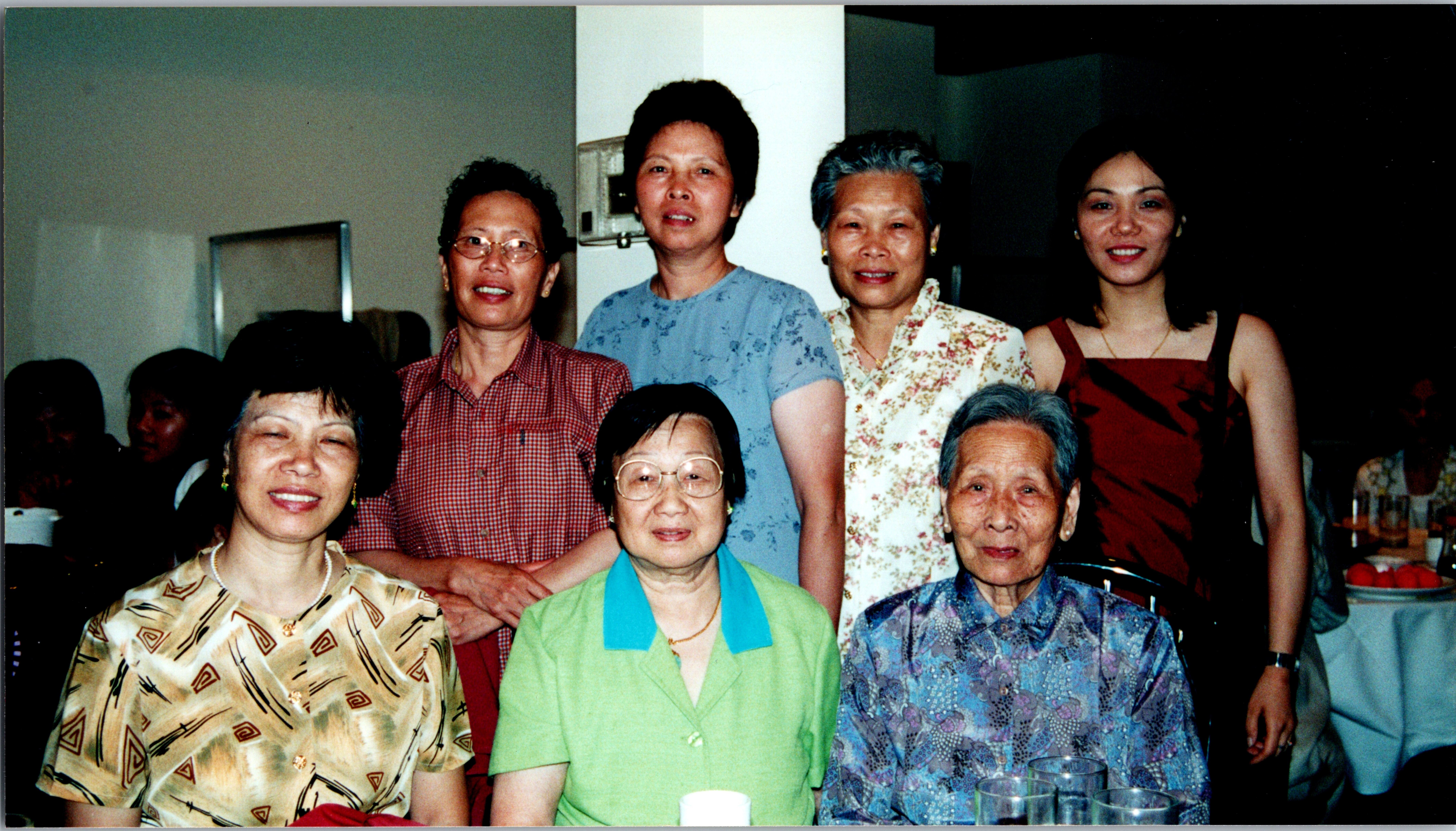 Group of elderly and middle-aged women posing together at a dinner party, smiling indoors at a long table with drinks in the foreground