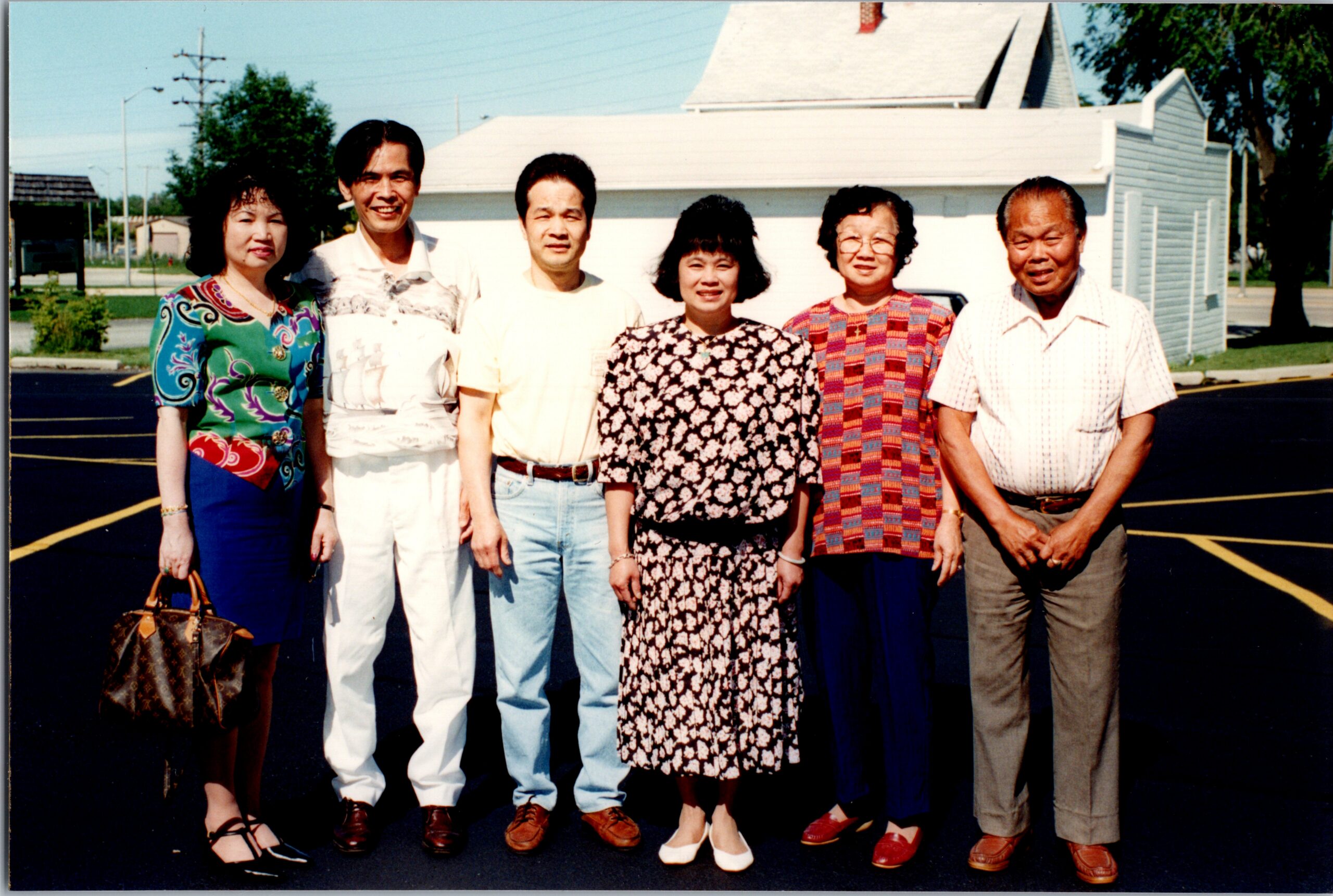 Seven adults standing side by side outdoors in a parking lot, posing for a group portrait in casual, colorful clothing.