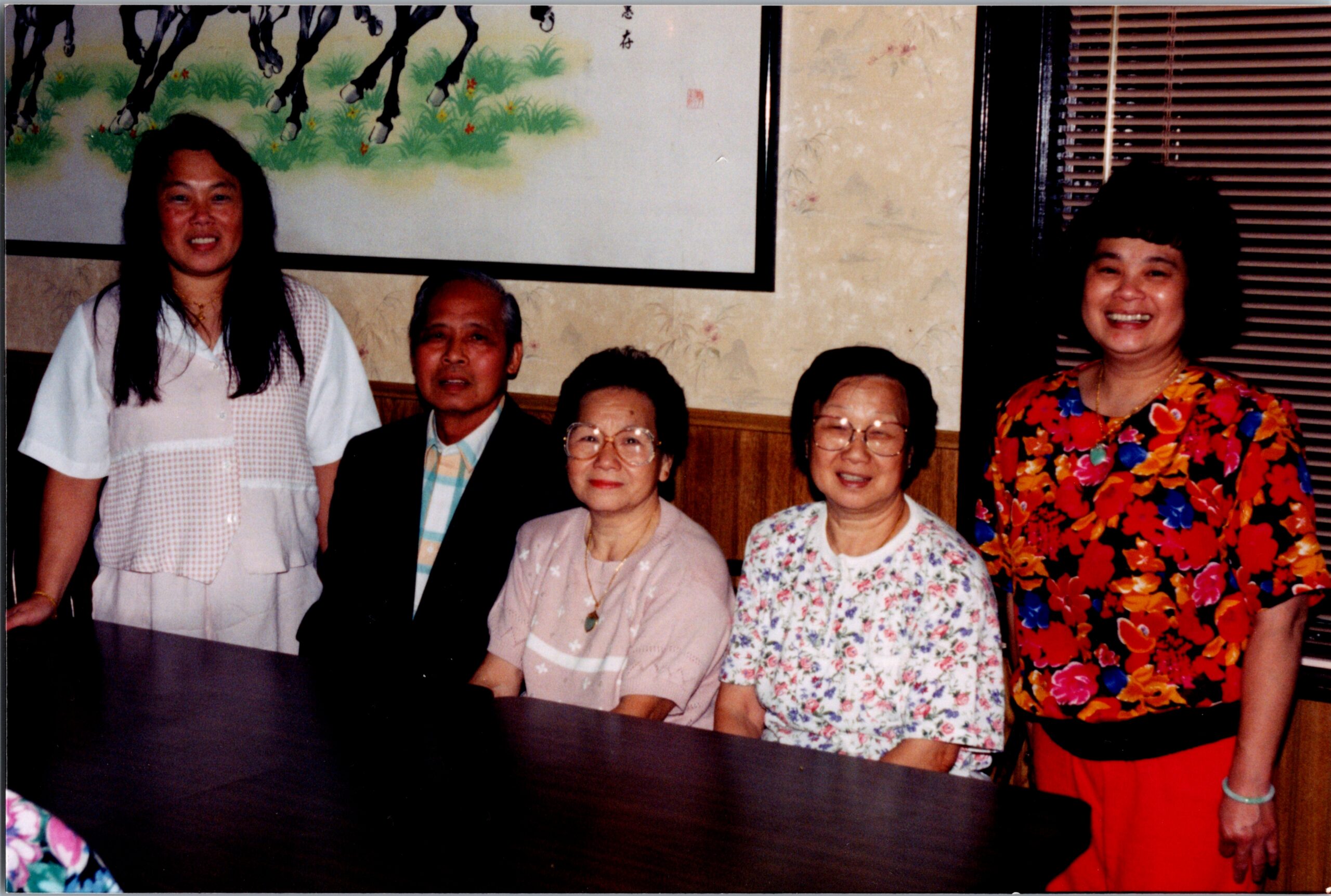 Five people pose for a group photo indoors: a woman in white on the left, a man and two elderly women seated in the middle, and a woman in a bright floral dress on the right, all smiling.