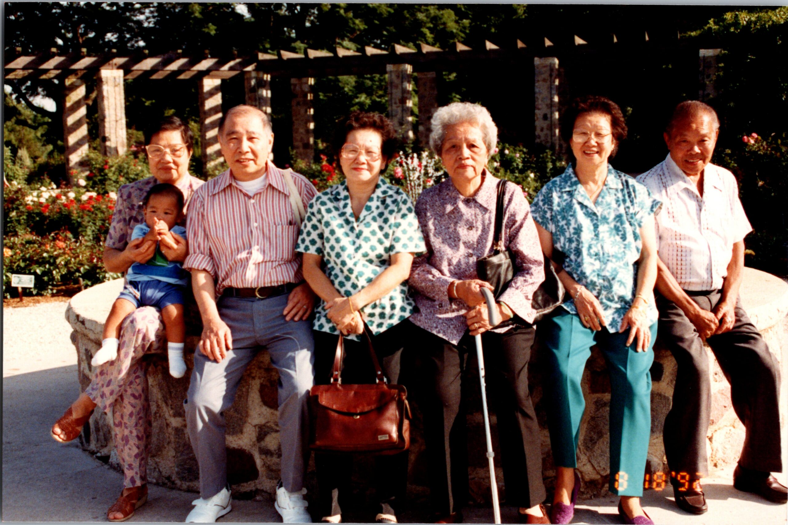 Group of seven seniors and a small child posing outdoors in a garden, smiling for the photo.