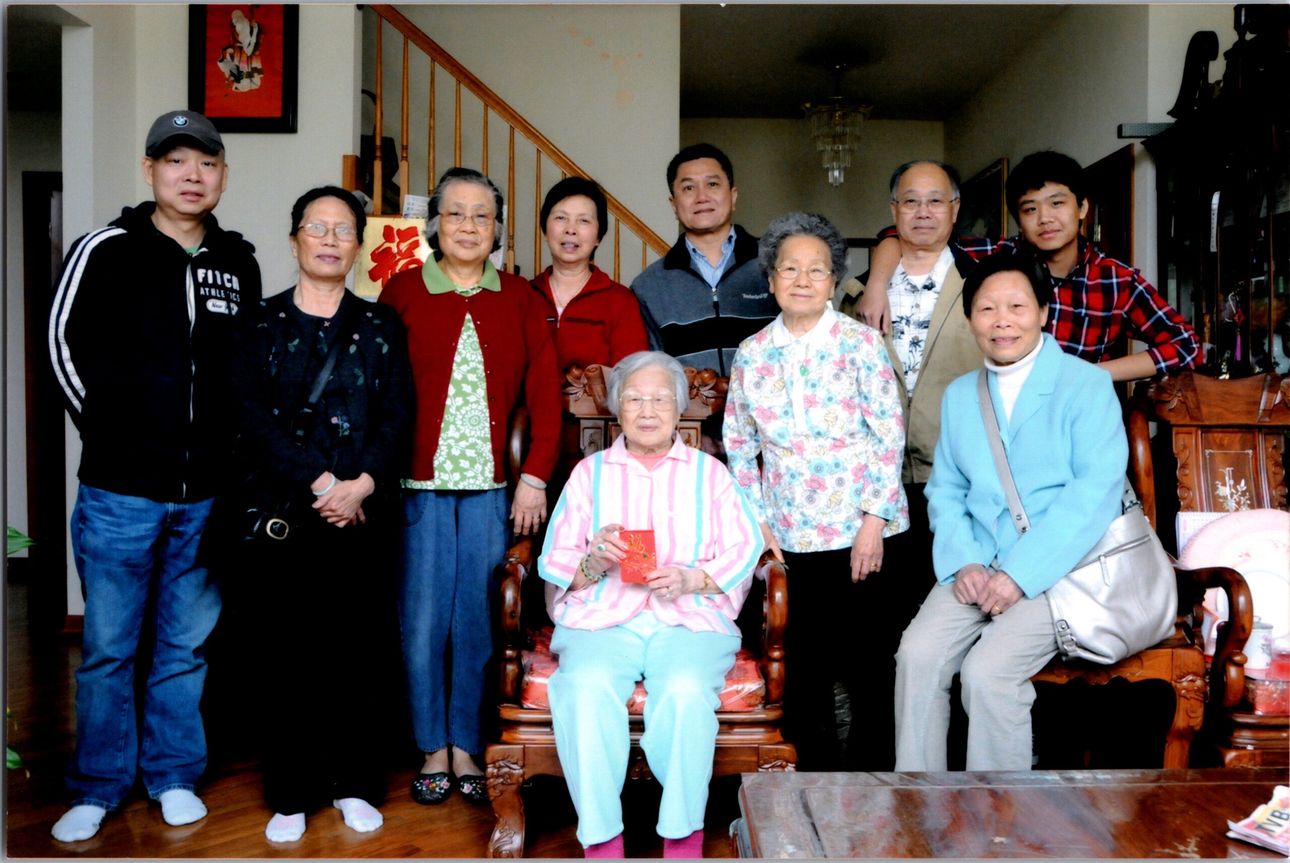 Group of ten Asian family members posing indoors for a group photo, two elderly women seated in front and others standing behind them, some holding red envelopes.