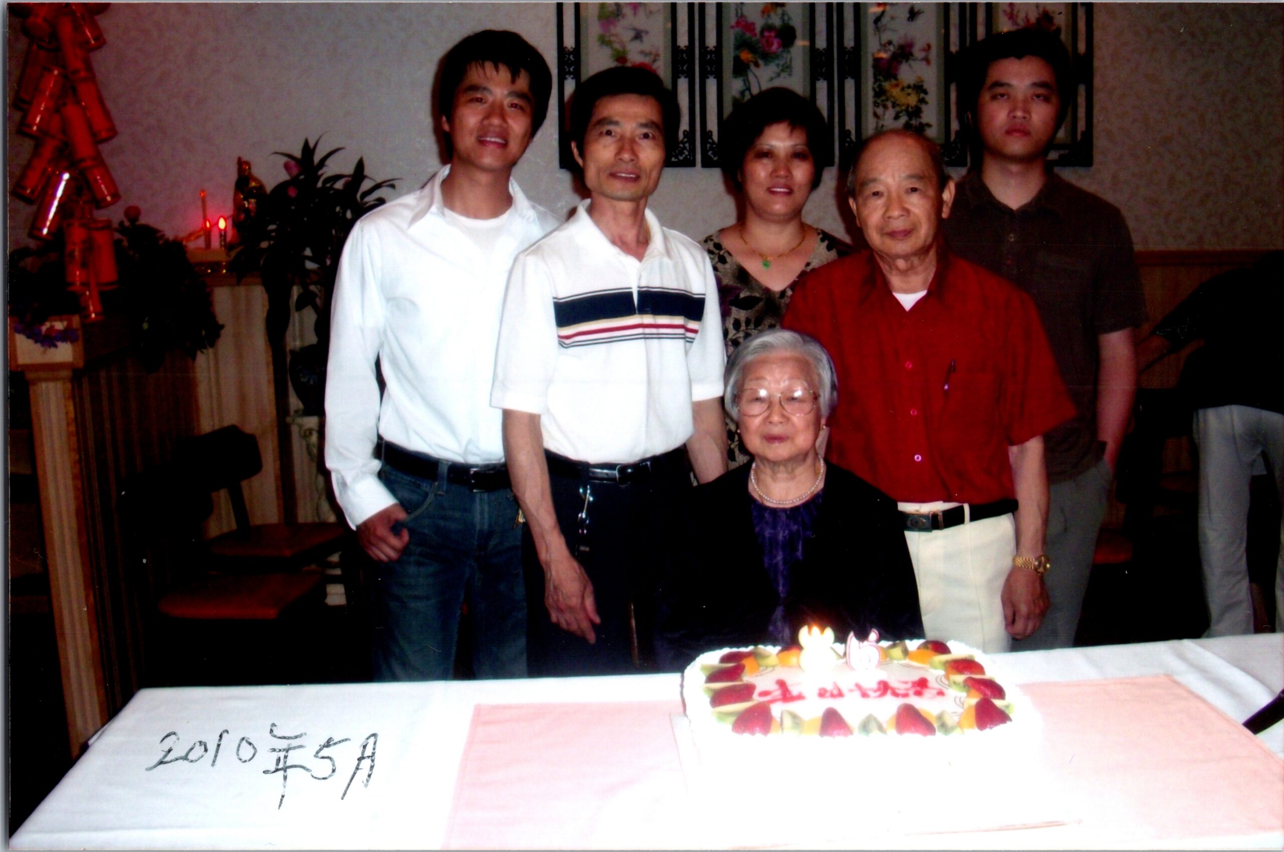 Group of relatives posing behind a birthday cake on a table; elderly woman seated at front, others standing around, date written in Chinese (2010年5月).