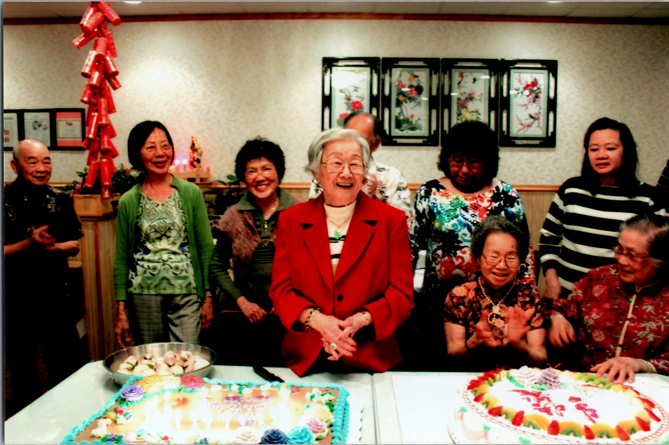Group of people at a cake party, smiling around two colorful decorated cakes on a table; central elderly woman in a red jacket leads the photo.