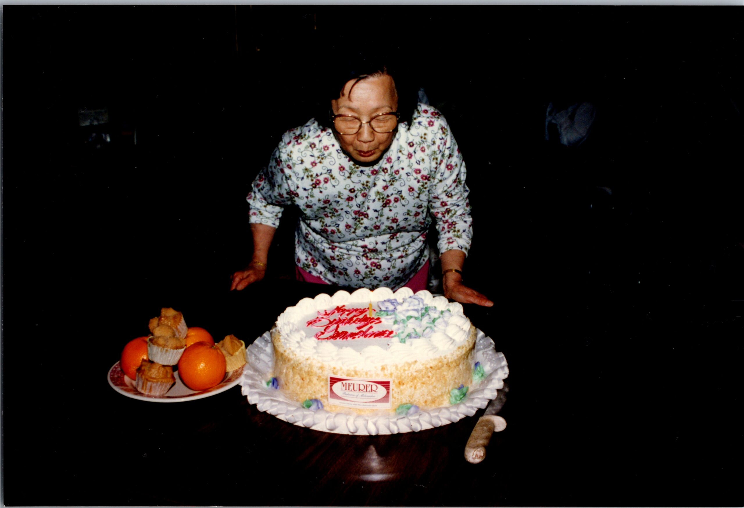 Older woman in a floral blouse leans over a two-layer birthday cake with candles, fruit and pastries nearby in a dark setting.