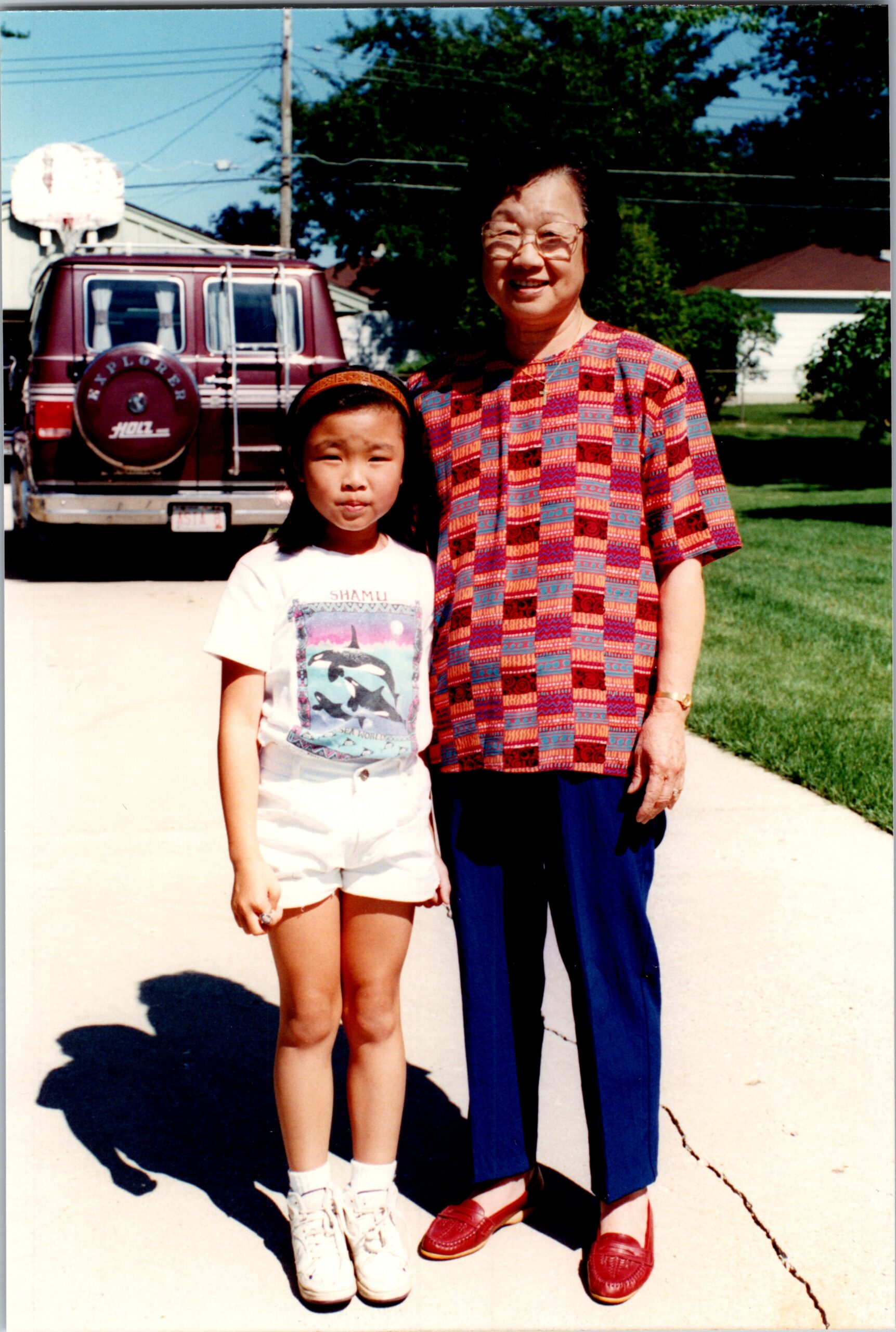 Grandmother and young girl posing together in a driveway with a maroon SUV in the background on a sunny day.