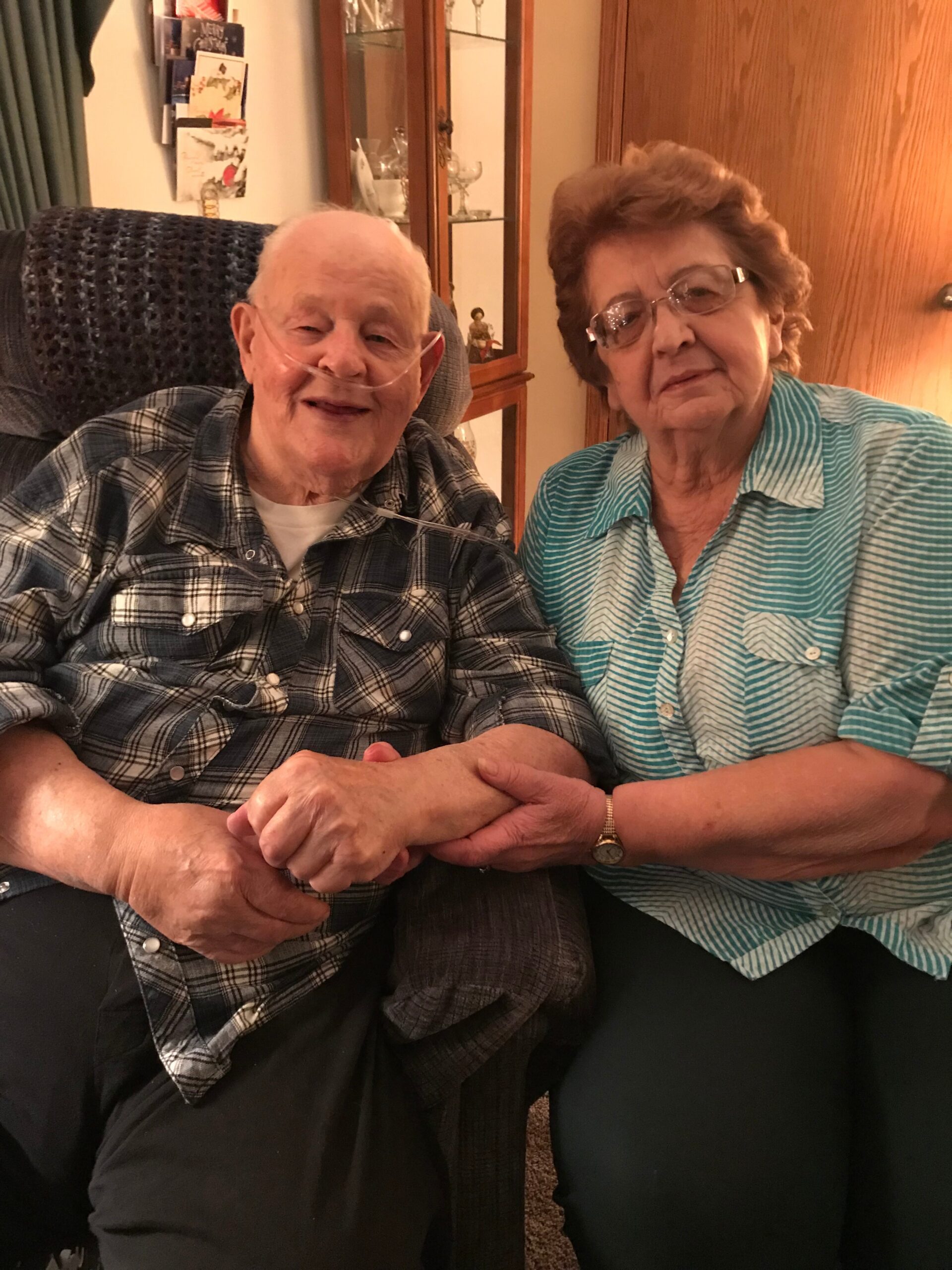 An elderly couple sits together on a couch, smiling; the man has a nasal oxygen tube and they hold hands.