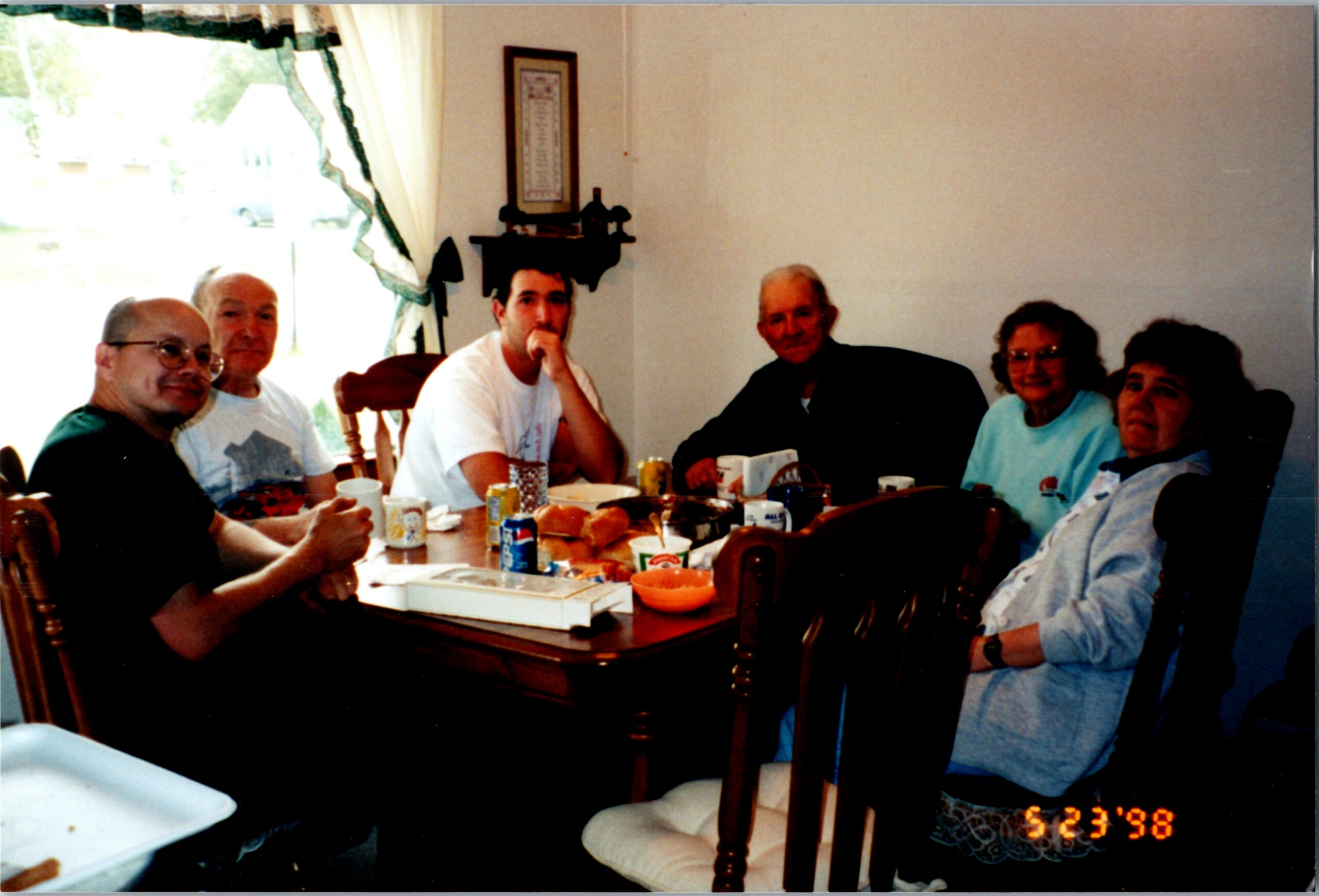 Group of six adults seated around a dining table with cups, snacks, and pastries in a home setting, looking at the camera.