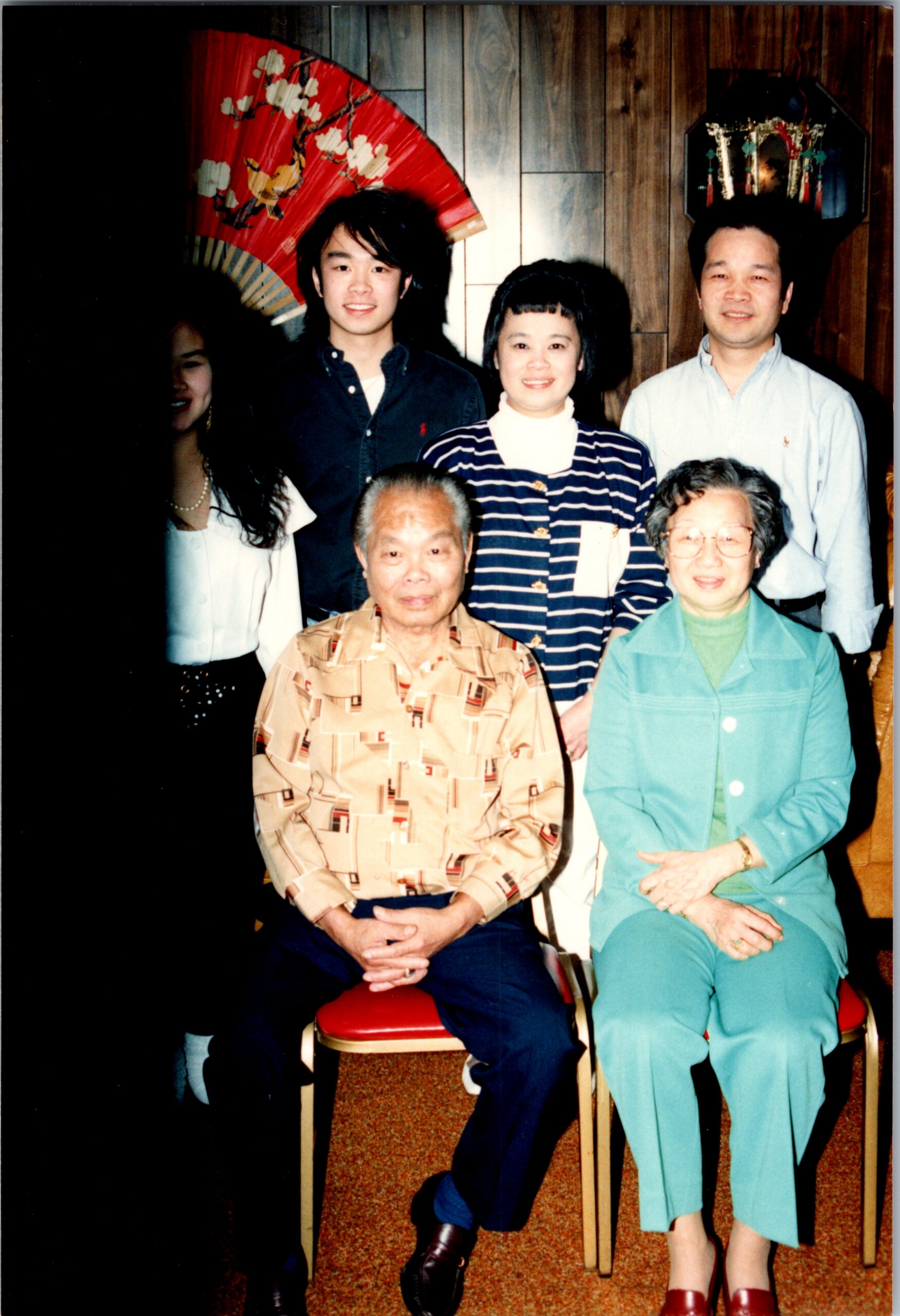 Group family portrait: two elderly seated in front, four adults standing behind, in a room with wood paneling and decorative items including a red fans and hair ornaments on display.