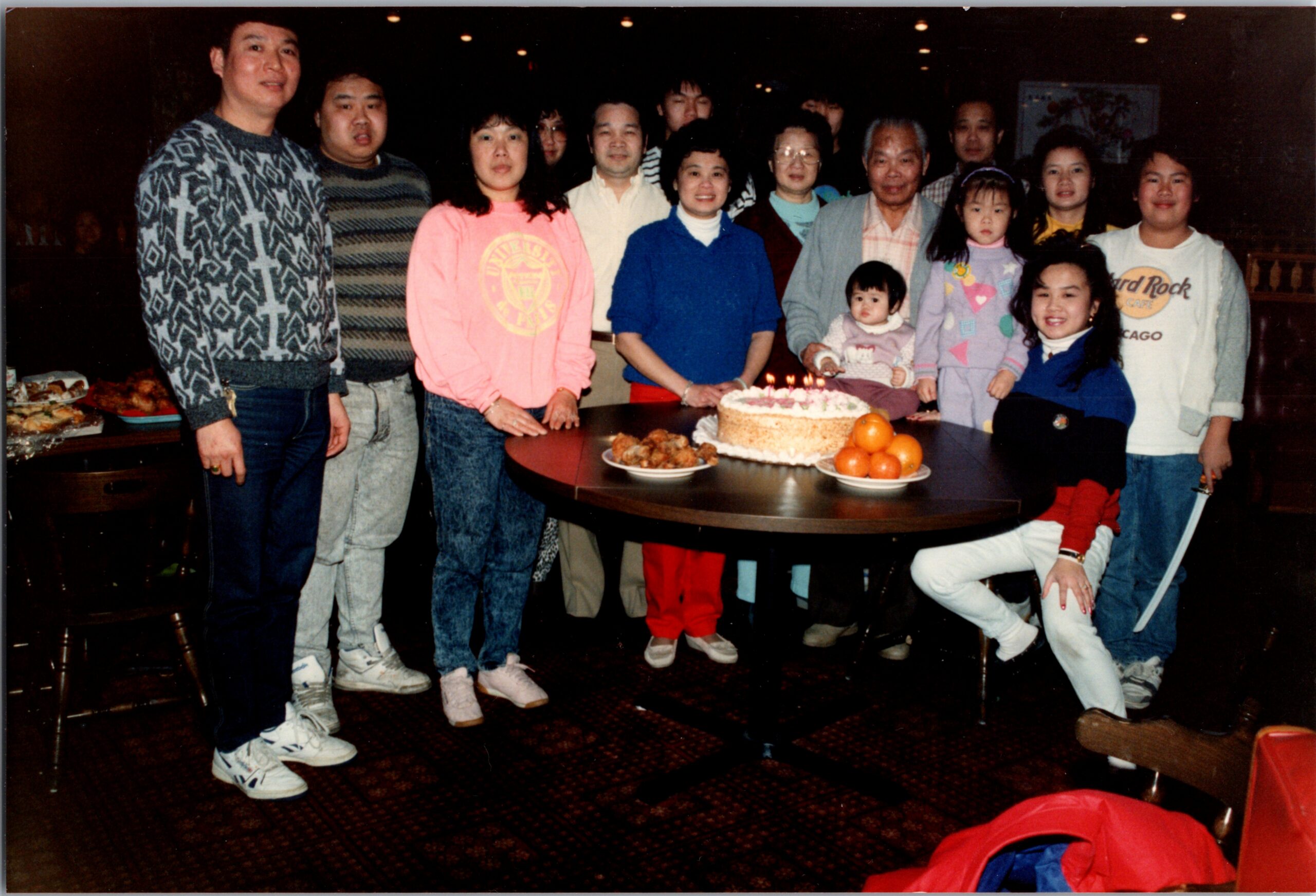 Group of family and friends posing behind a birthday cake with lit candles on a table, with plates of snacks and oranges nearby in a dimly lit room.