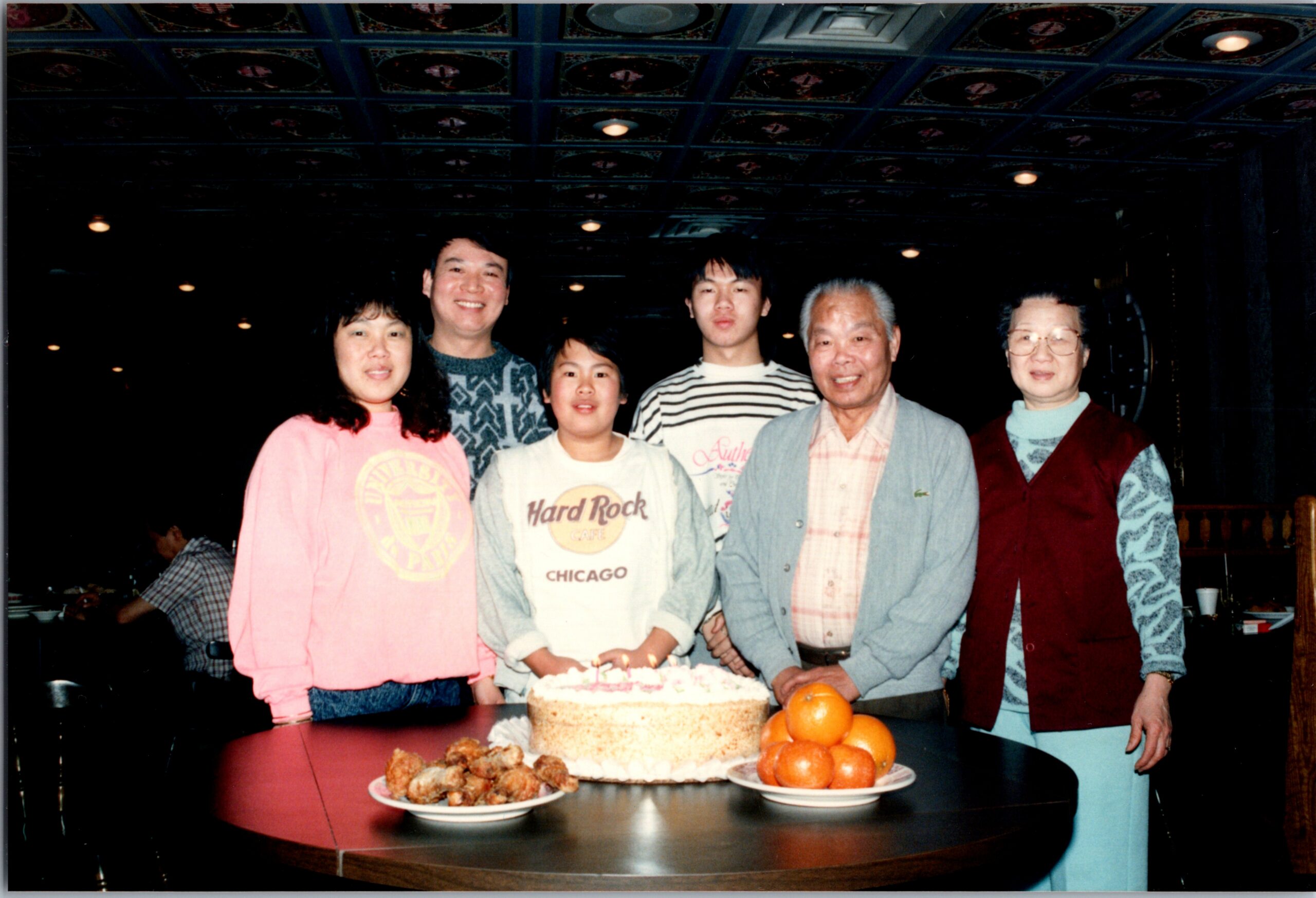 Six people—young adults and seniors—pose around a birthday cake on a restaurant table, with oranges in the foreground.