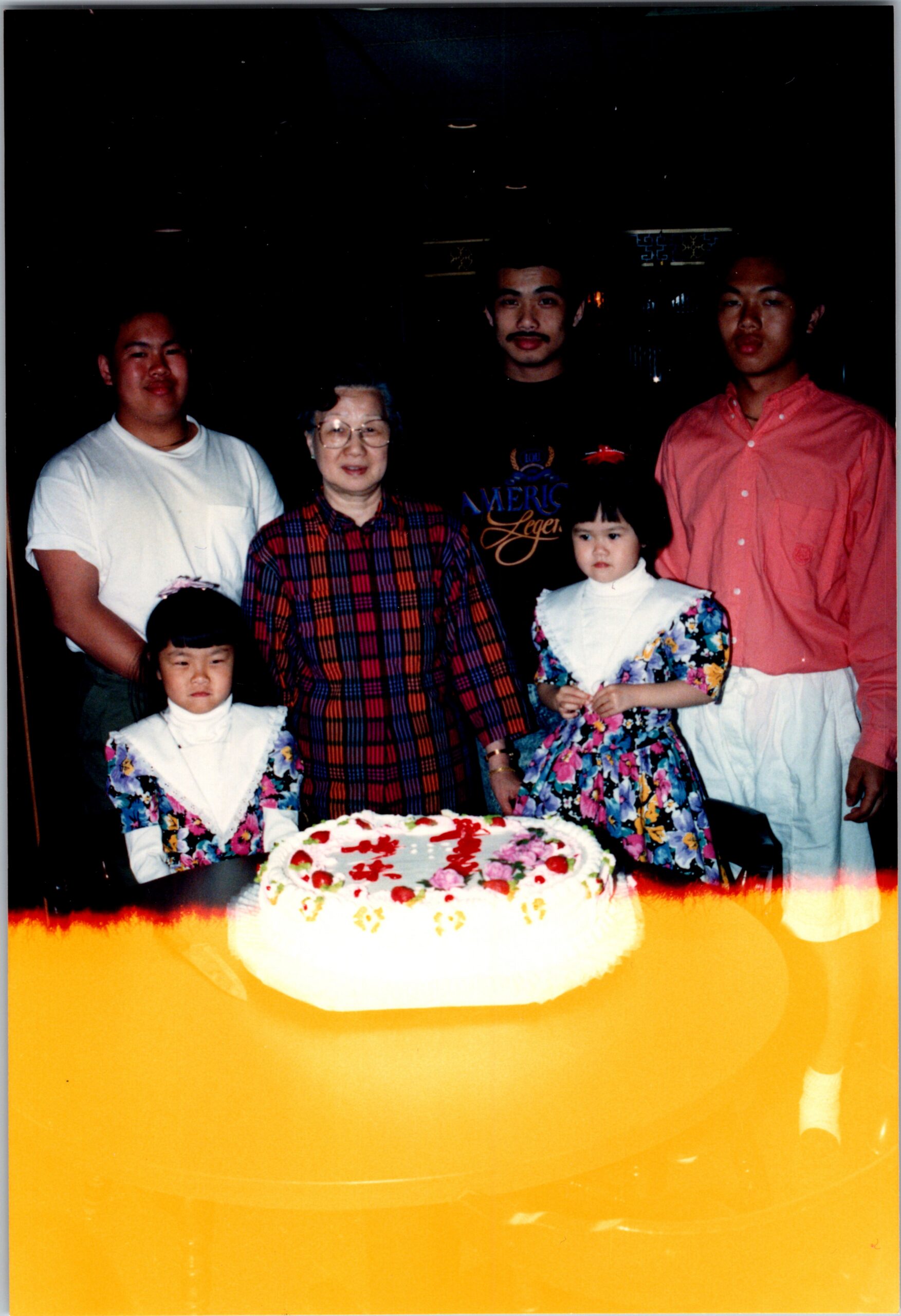 Family birthday photo: six people posing behind a decorated cake, including an elderly woman, two girls in floral dresses, and three men in casual clothes.