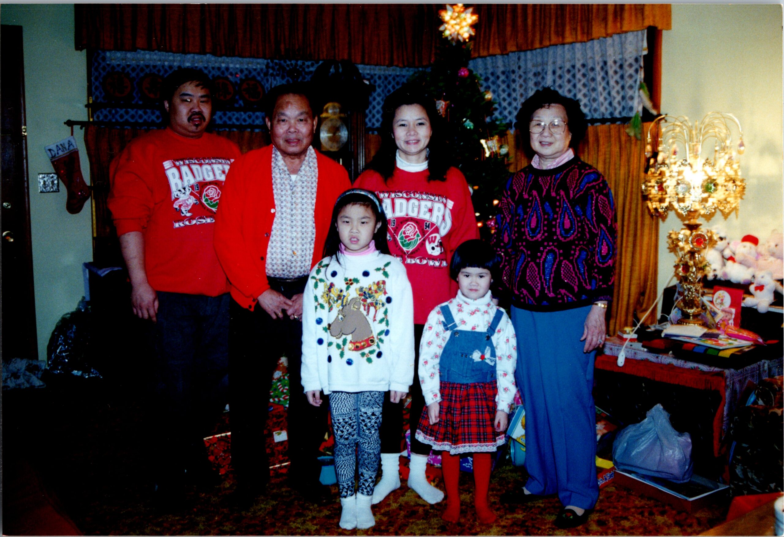 A family of six posing for a Christmas photo in a living room with a decorated tree and gifts nearby.