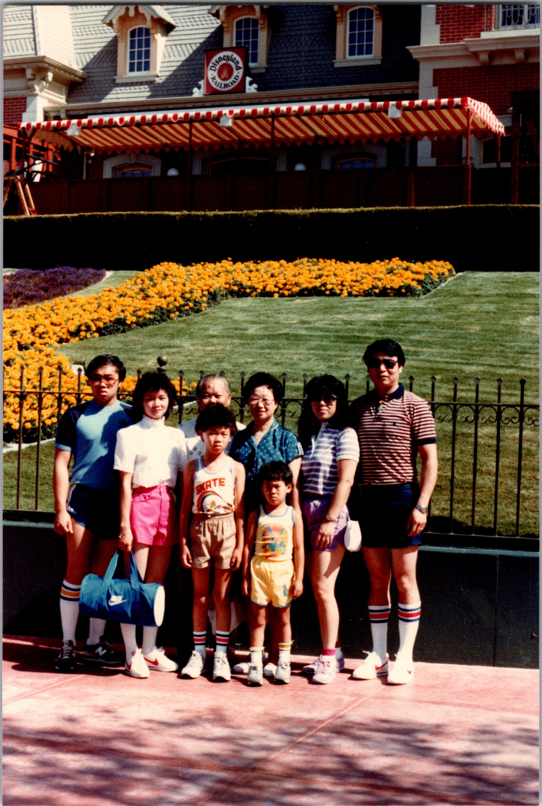Family group photo in front of a Disneyland Railroad building with a red-and-white striped canopy, orange flower bed, and well-manicured lawn in the background.