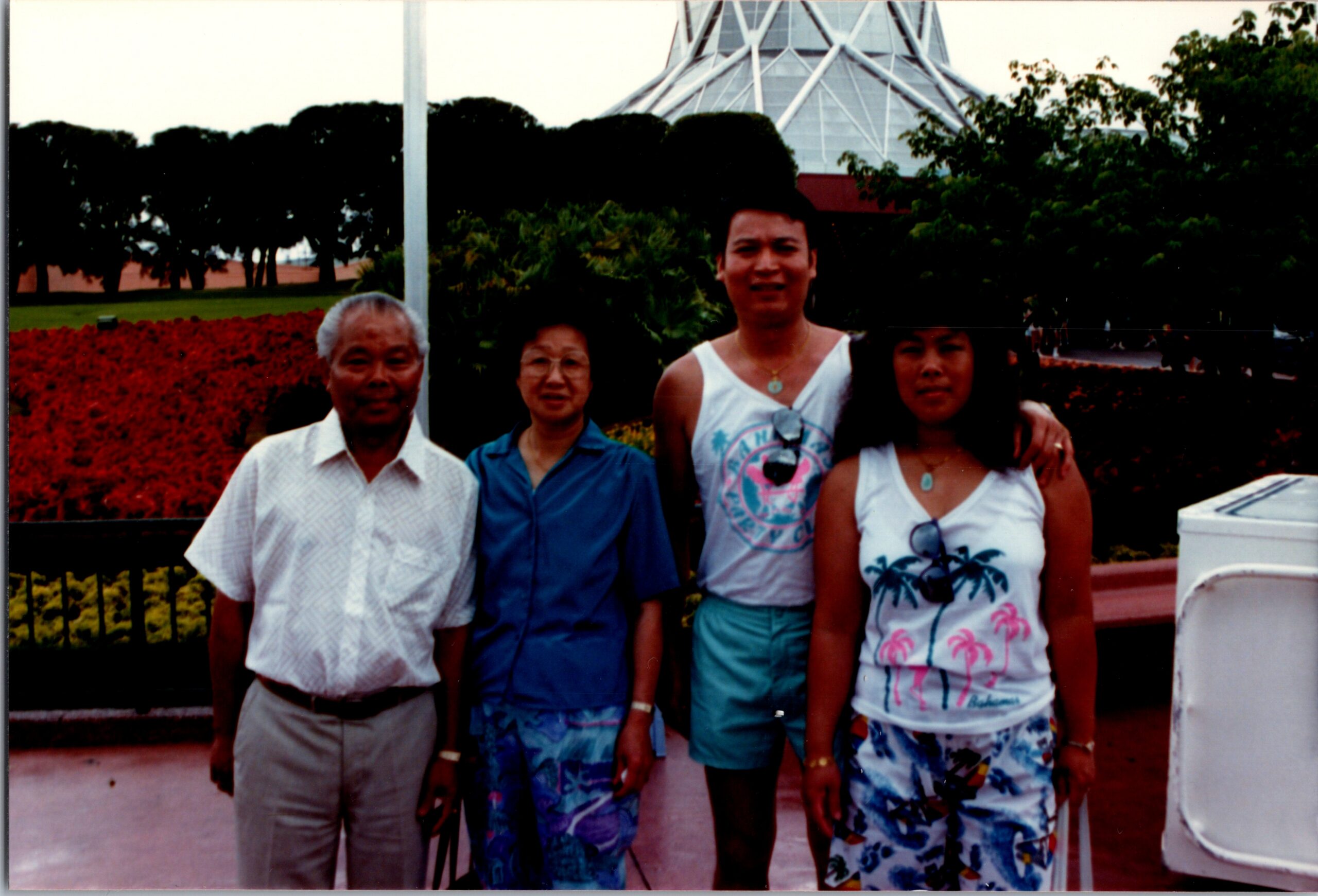 Four people stand together outdoors in front of a vibrant red flower bed and a modern glass pavilion in the background.