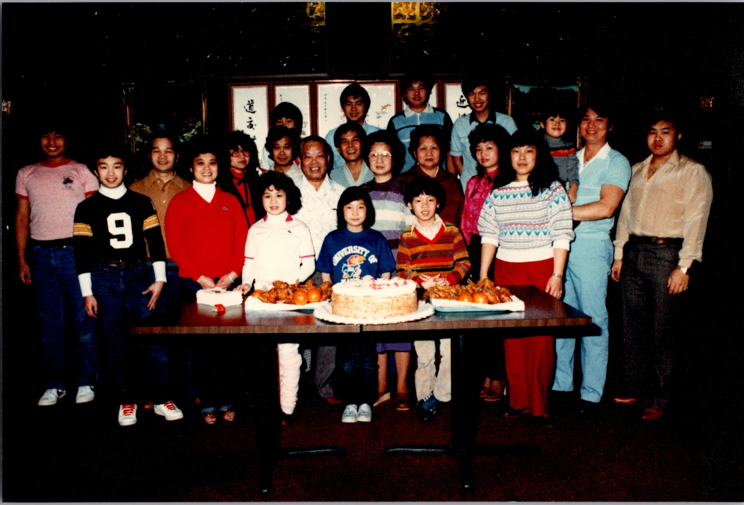 Group of adults and children posing behind a table with a birthday cake and fruit on display; dimly lit room.
