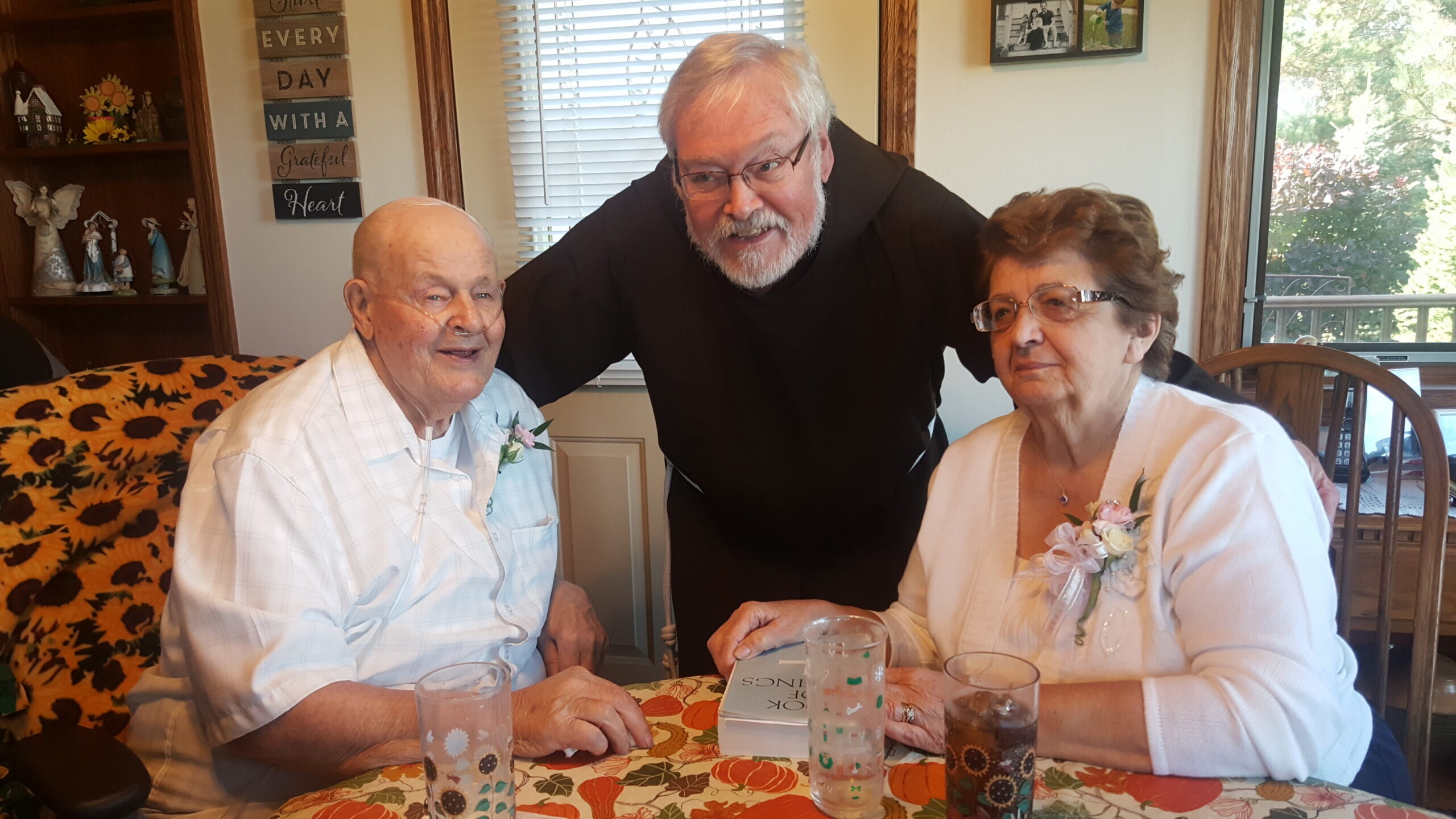 Three elderly people sit around a floral tablecloth, with a man in a black shirt leaning between them in a warm home gathering.