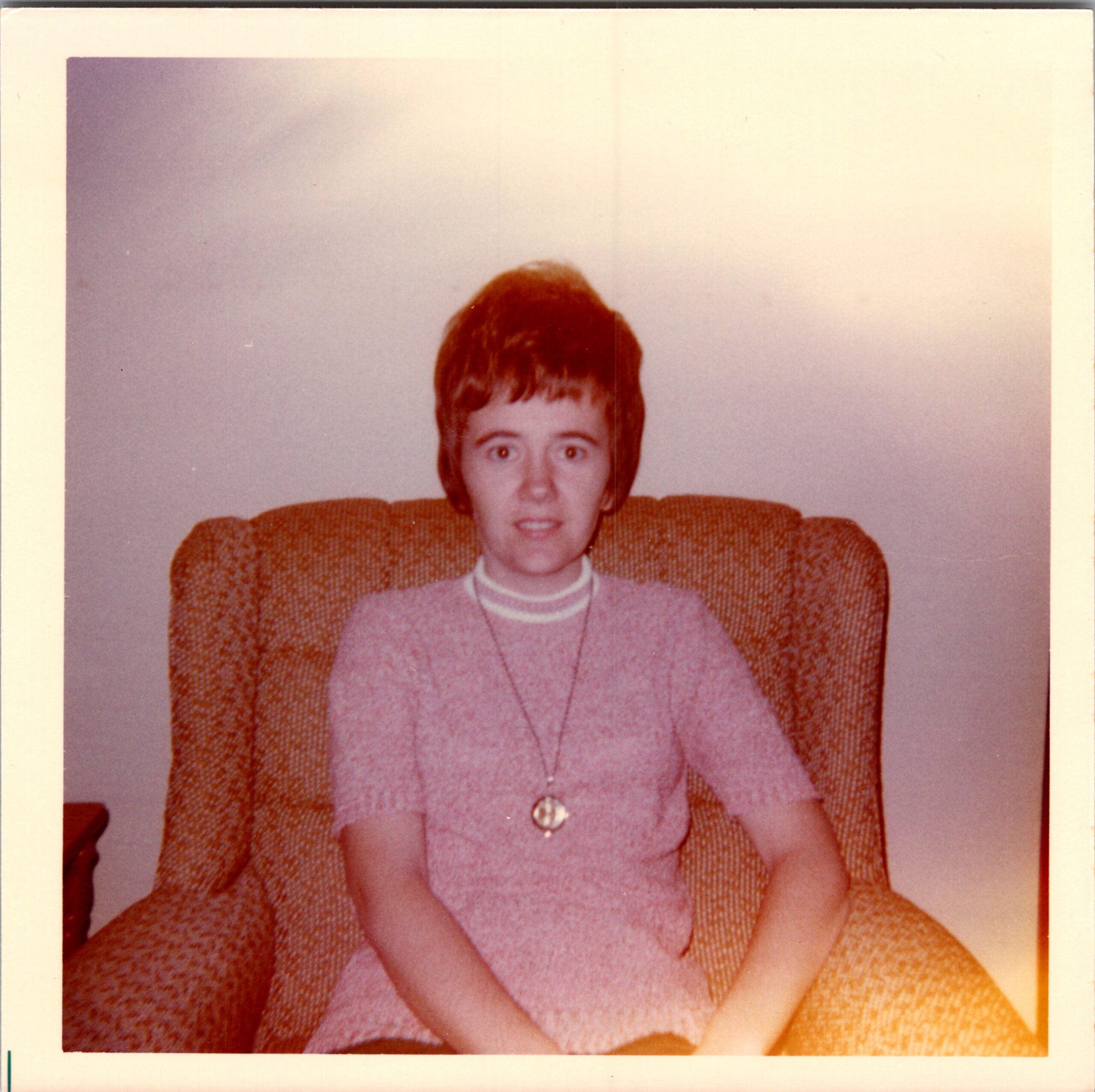 Portrait of a woman with short hair wearing a pink sweater and necklace, seated on an orange couch, looking at the camera.