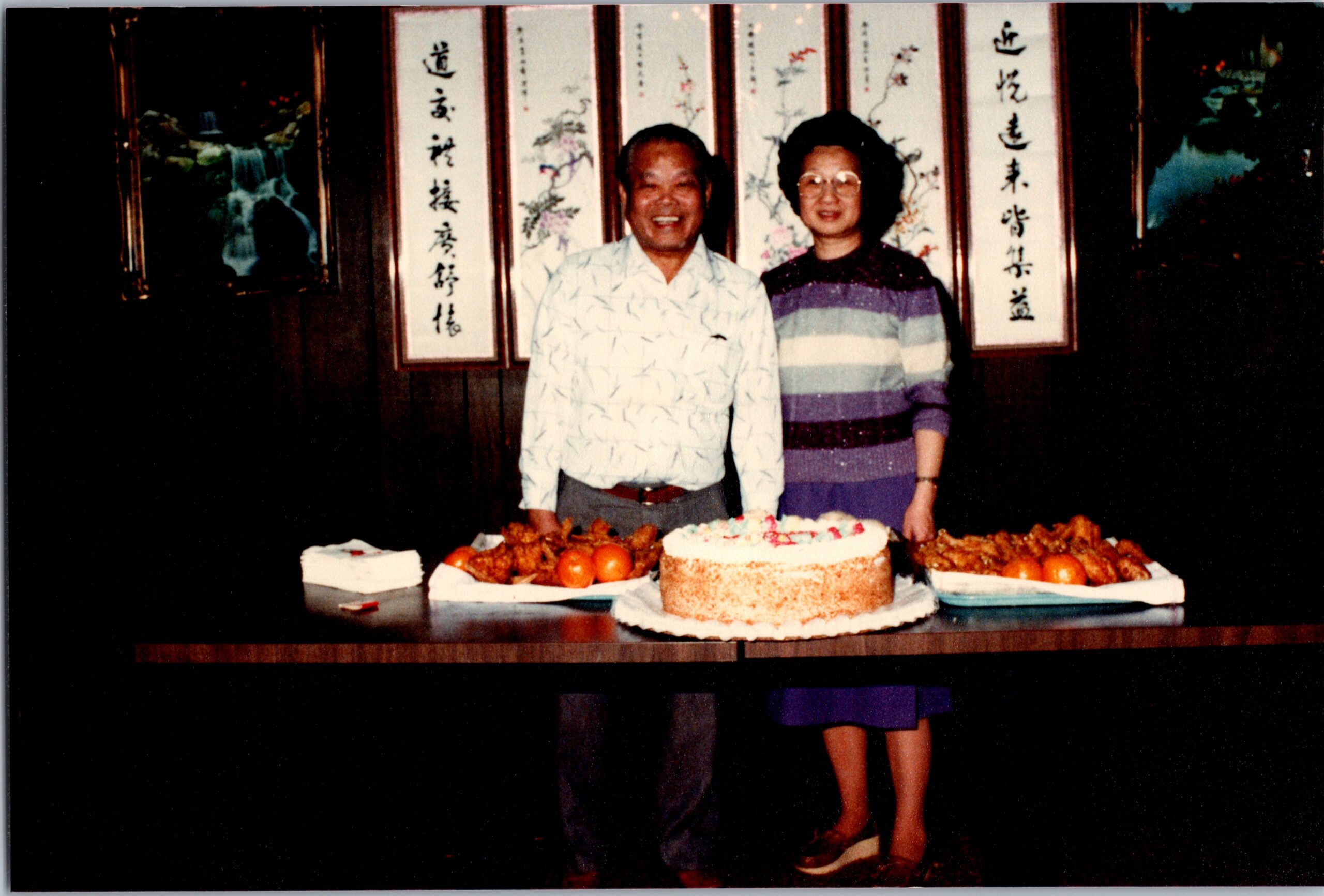 Two adults smile beside a large frosted cake and bowls of fruit on a table, with decorative scrolls in the background of a celebration setting.