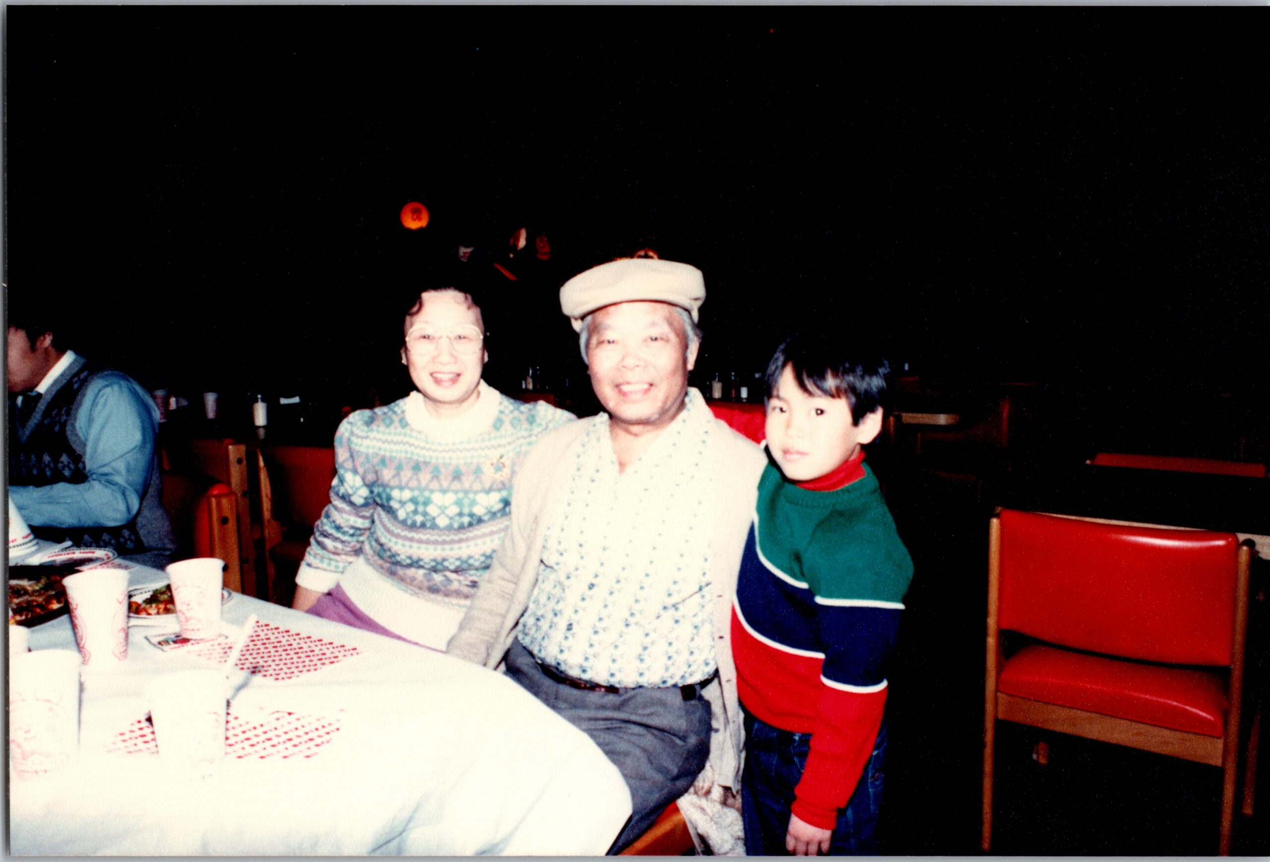 Three people pose at a holiday dinner table: a smiling woman in glasses, an older man in a hat, and a child in a red and green sweater with cups and plates nearby.