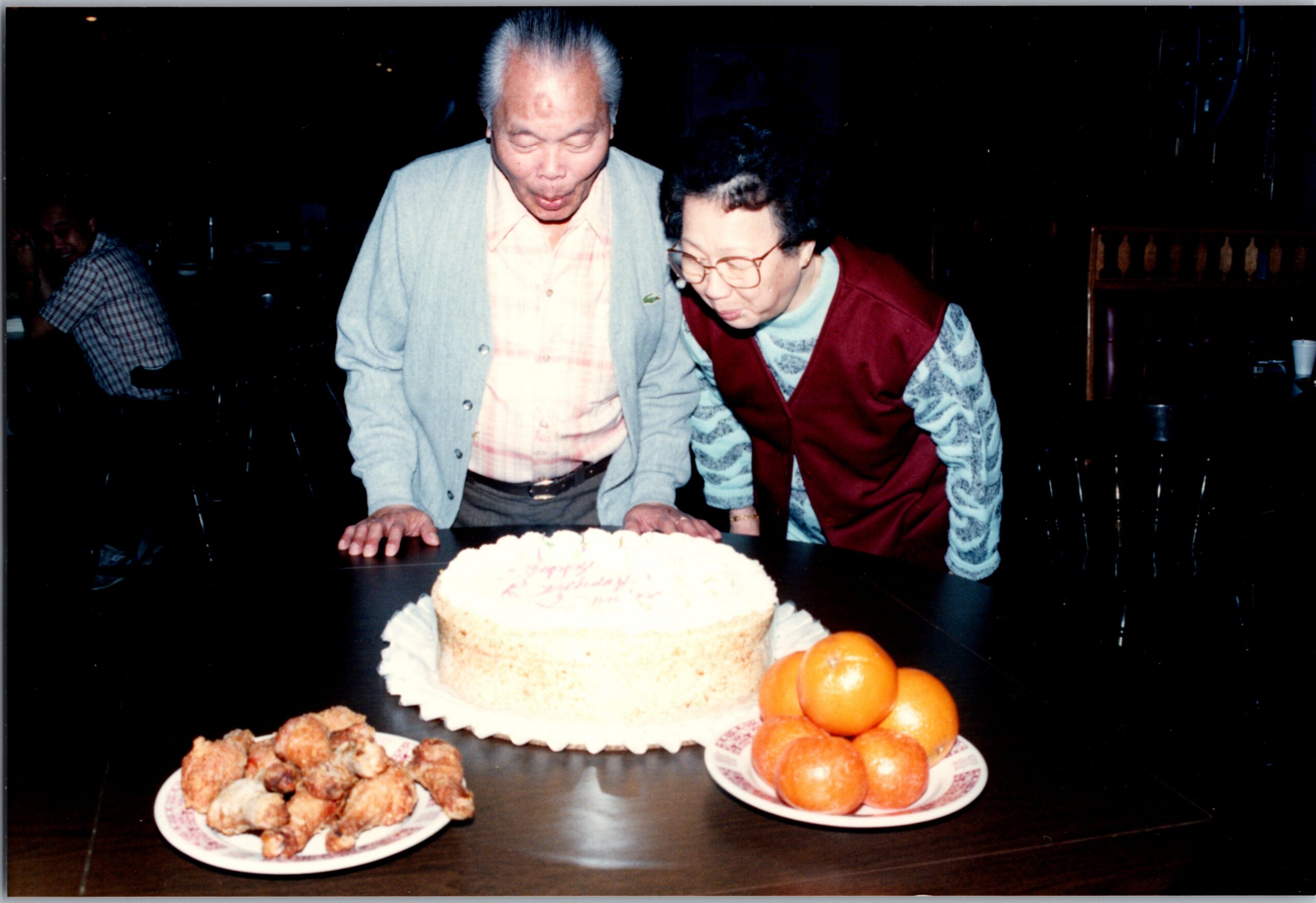 Two elderly people lean over a birthday cake on a table, about to cut it, with plates of oranges and fried snacks nearby in a dim restaurant setting.
