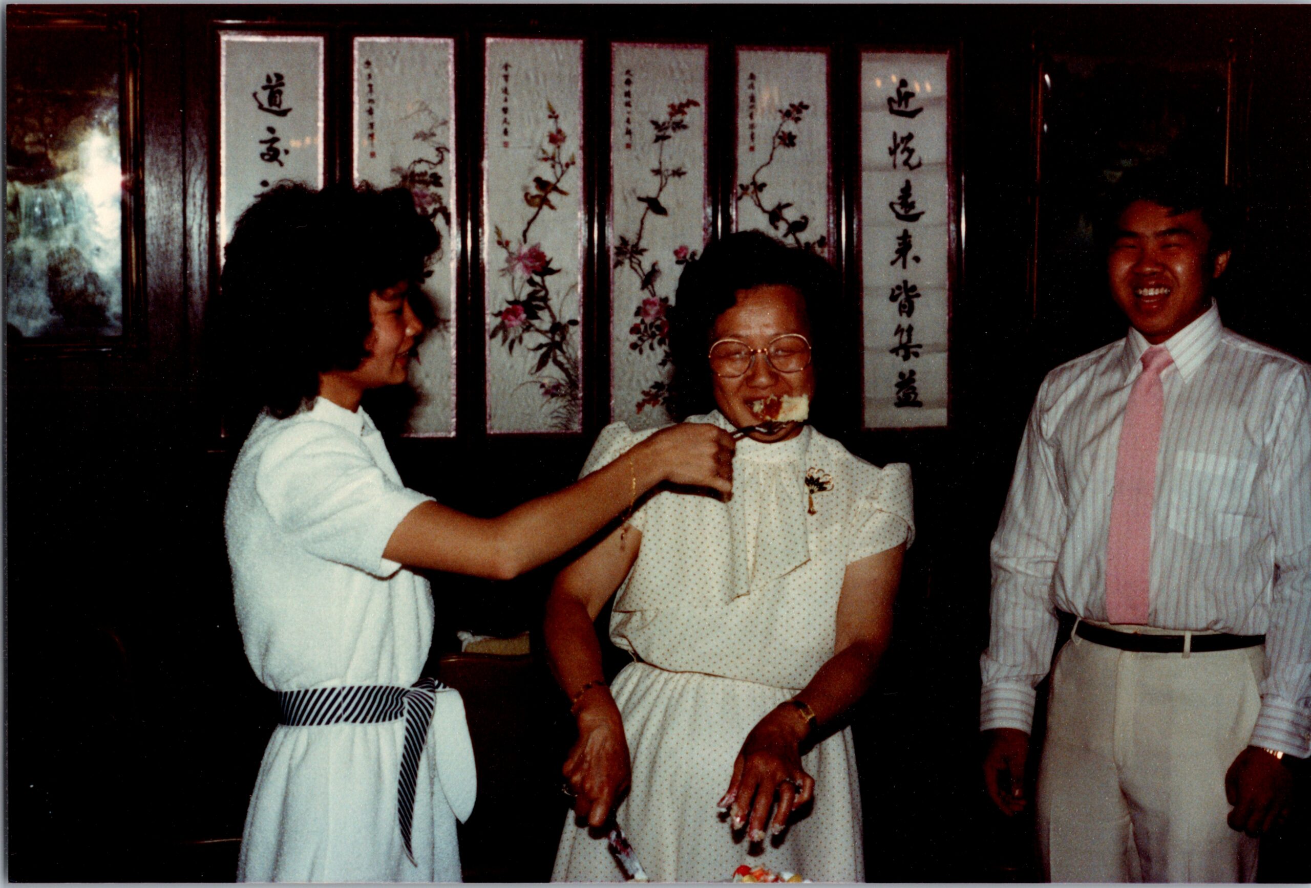 Older woman in a polka-dot dress is fed cake by two friends at a social gathering, smiling and enjoying the moment, with decorative screens in the background.