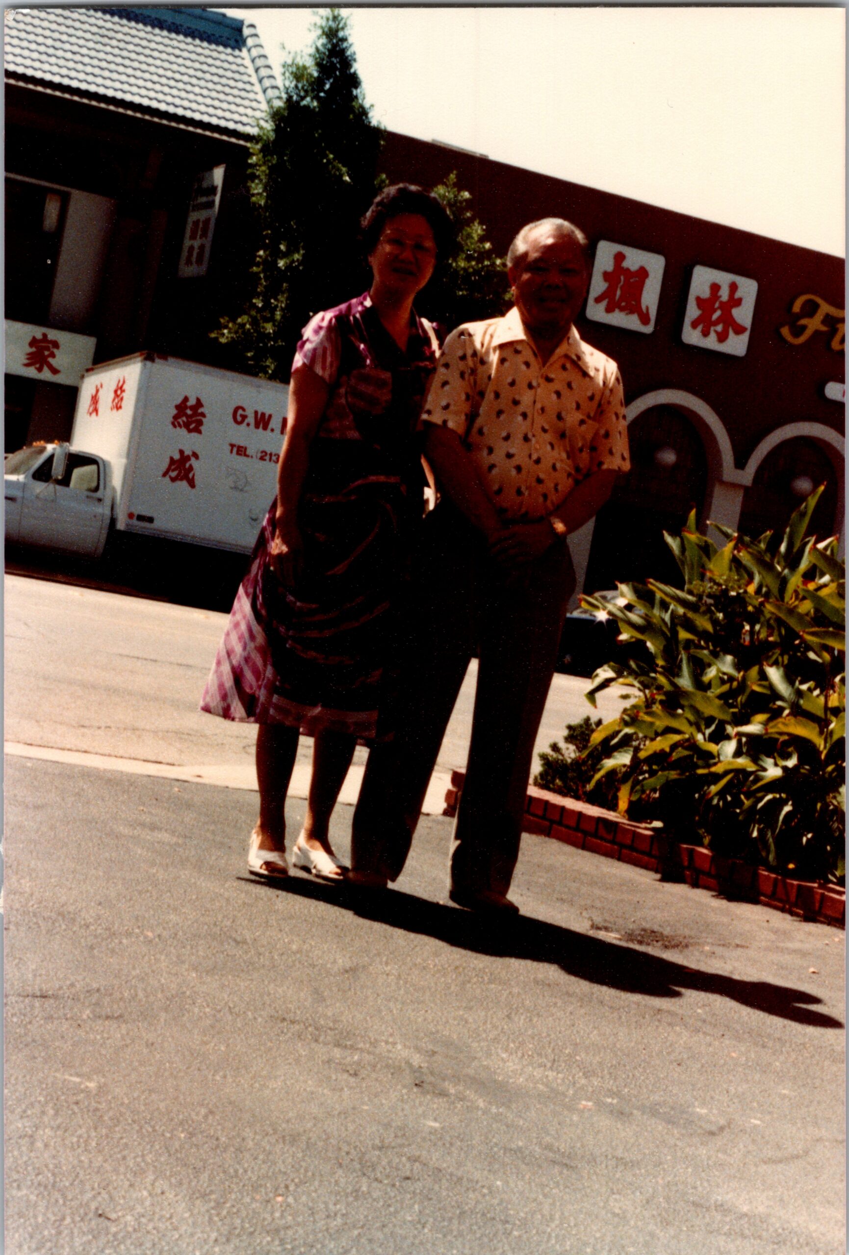 Older couple posing on a sidewalk in front of a building with Chinese signboards behind them.