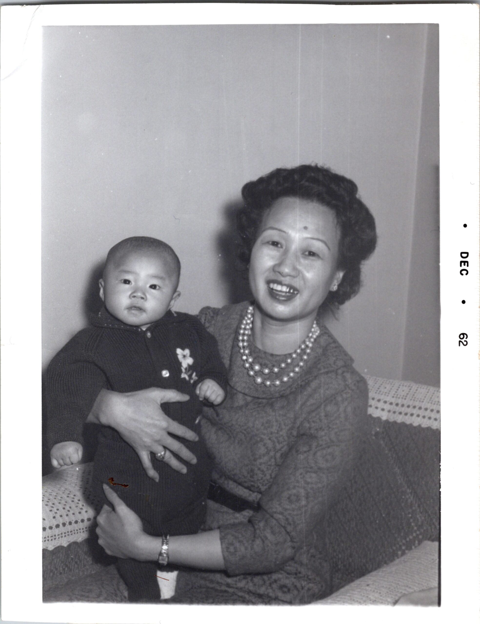 Smiling woman in a textured dress and multiple pearl necklaces holding a baby indoors, both looking at the camera (black-and-white photo).
