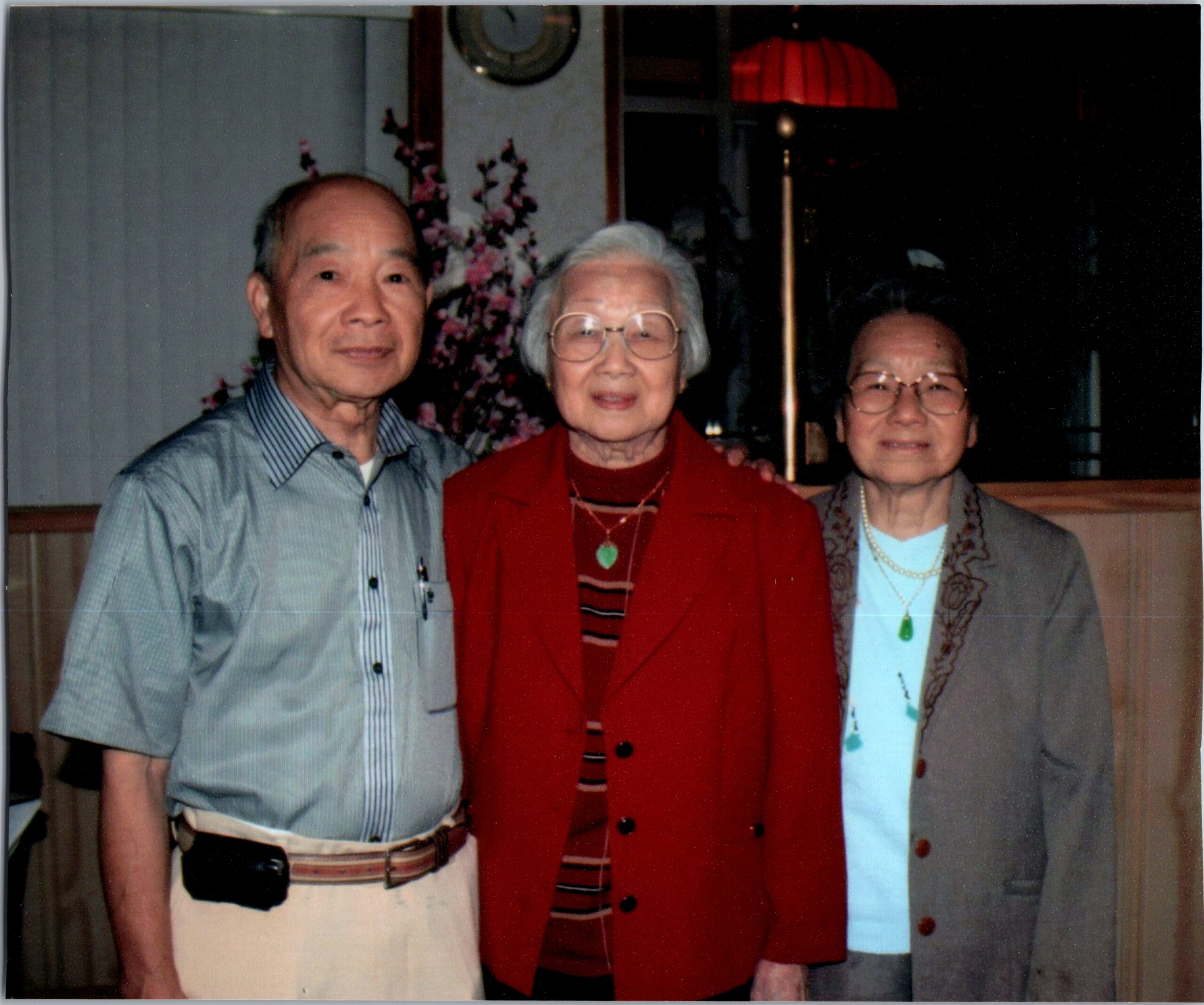 Three elderly adults posing indoors: a man in a striped shirt on the left, a woman in a red blazer in the center, and a woman in a gray cardigan on the right; floral arrangement and a red lamp in the background.