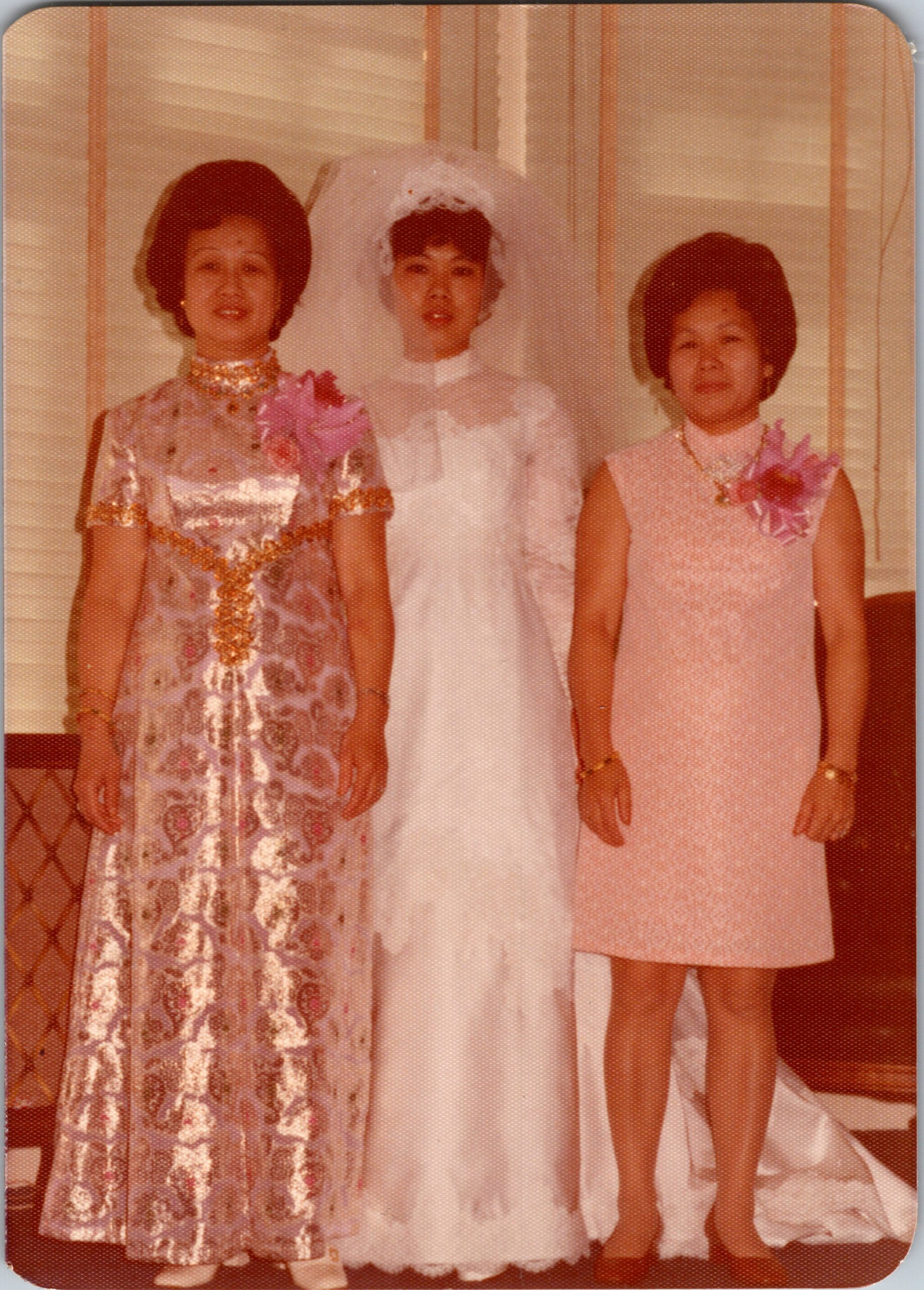 Three young girls in formal dresses posing together for a photo indoors, two wear pink, one in white with a veil and flowers.