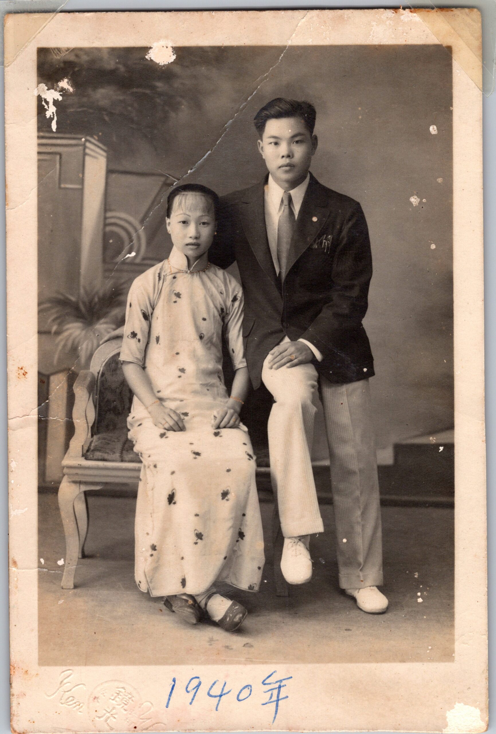 Portrait of a young Asian couple in a studio setting: woman seated in a floral cheongsam, man standing beside her in a dark suit and tie; '1940' handwritten at bottom.